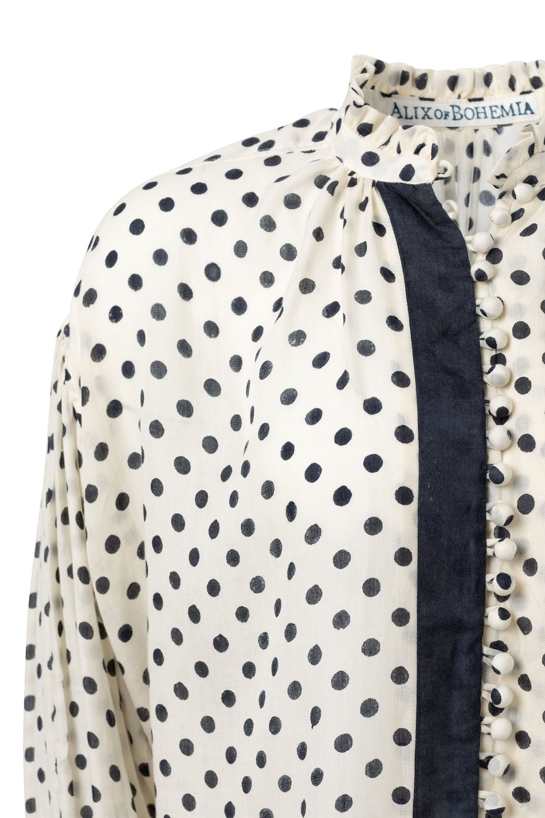 Close-up of the Prima Tuxedo Spot Blouse, featuring a white fabric with black polka dots and a dark vertical stripe. It is adorned with fabric-covered buttons and a slightly ruffled collar. Crafted from hand-printed Cotton Tencel, the label inside reads "Alix of Bohemia.