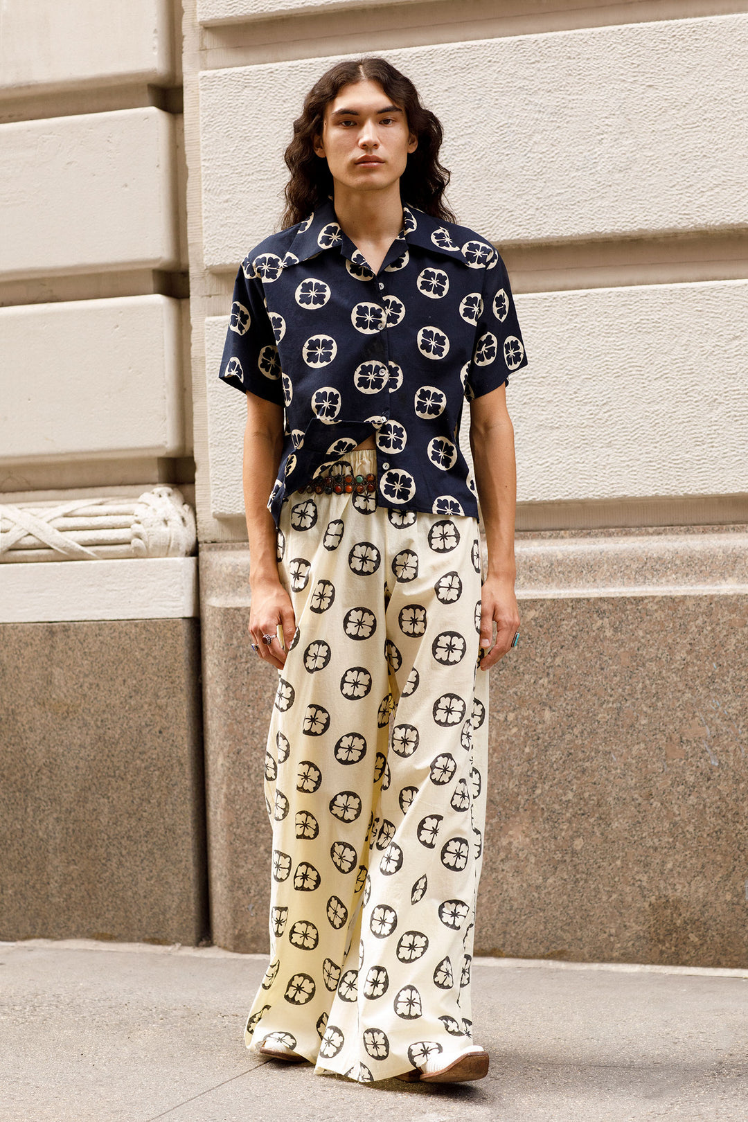 A person with long hair stands before a beige wall, radiating unique beauty in a Stevie Blue Iris Shirt and cream pants, both featuring black and white circular patterns. Their attire captures the vintage charm of a Hawaiian shirt, as the decorative wall elements frame the scene against the visible sidewalk.