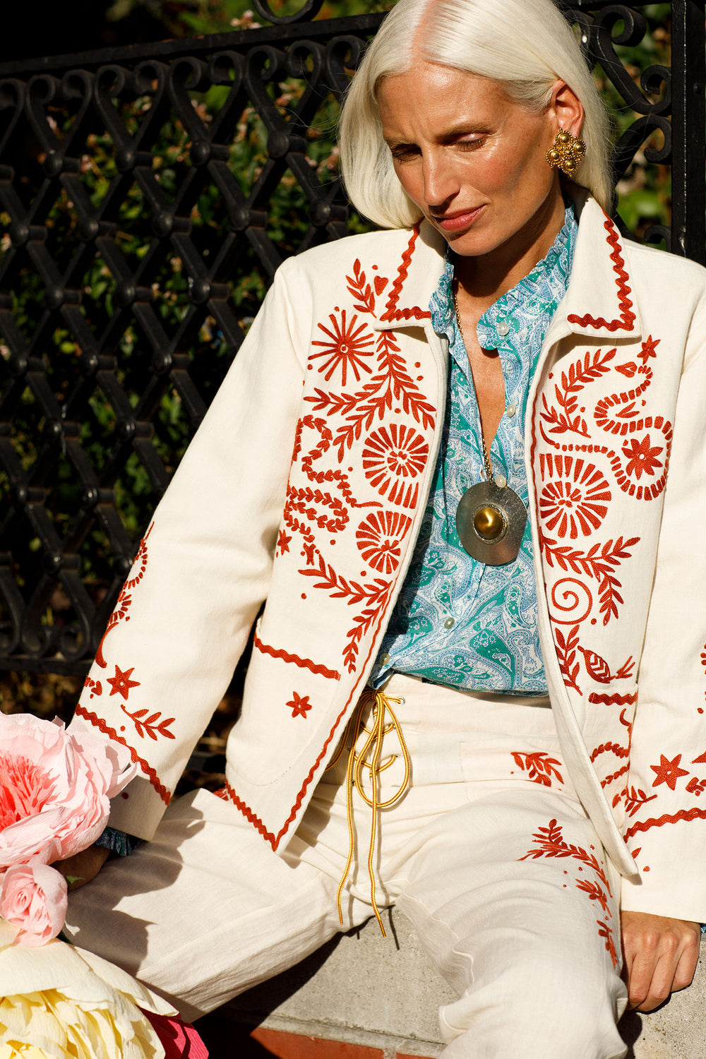 A person with long white hair sits outdoors, wearing a stylish white suit adorned with red floral embroidery and paired with a blue patterned shirt. The outfit, featuring hand-crafted intricate details, is complemented by the Penny Reef Pant, large round pendant, and gold earrings amidst pink flowers.