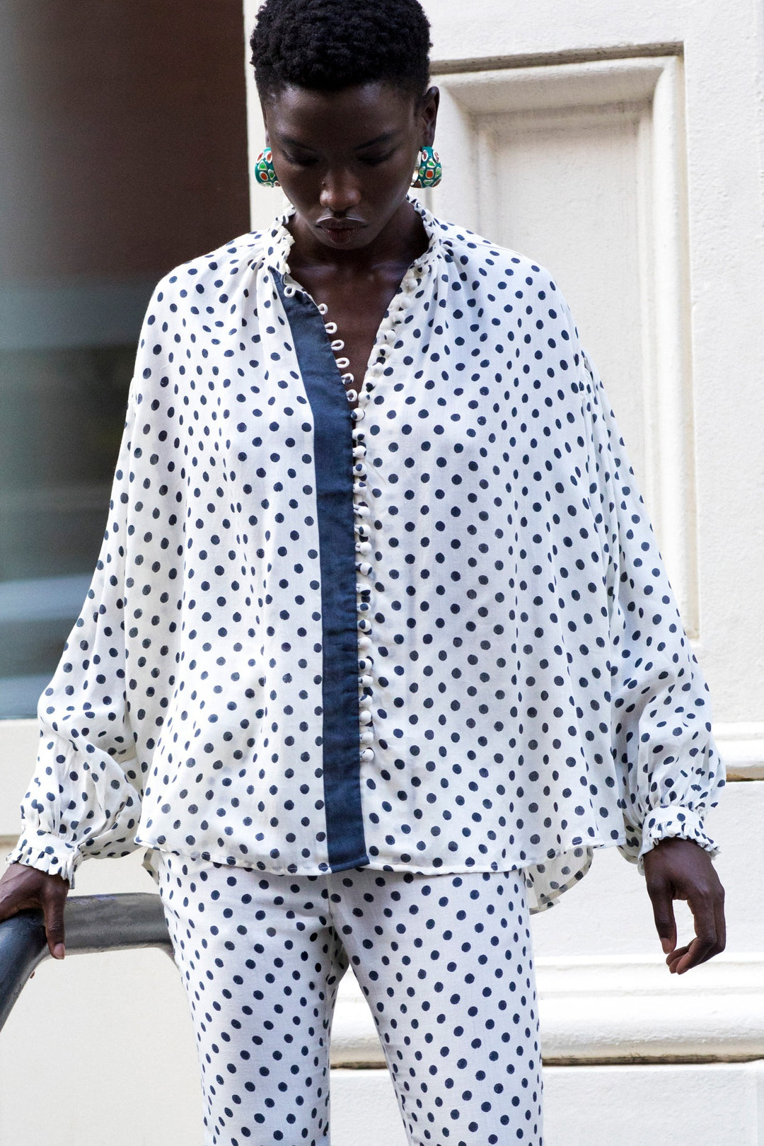 A person with short hair is standing near a light-colored wall, dressed in the Prima Tuxedo Spot Blouse, which is a loose-fitting white button-up adorned with black polka dots and featuring a ruffled collar, paired with matching Cotton Tencel pants.