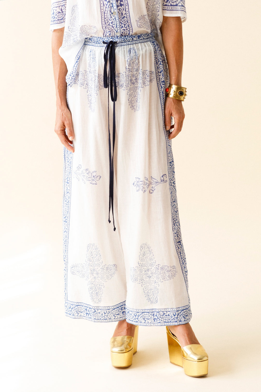 A person wearing the Natalie Criss Cross Pant ensemble, crafted from white cotton voile and adorned with blue hand block printed patterns, stands against a plain background. The outfit includes a flowing top and wide-leg pants featuring intricate designs. Gold platform shoes and a chunky bracelet complete the look.