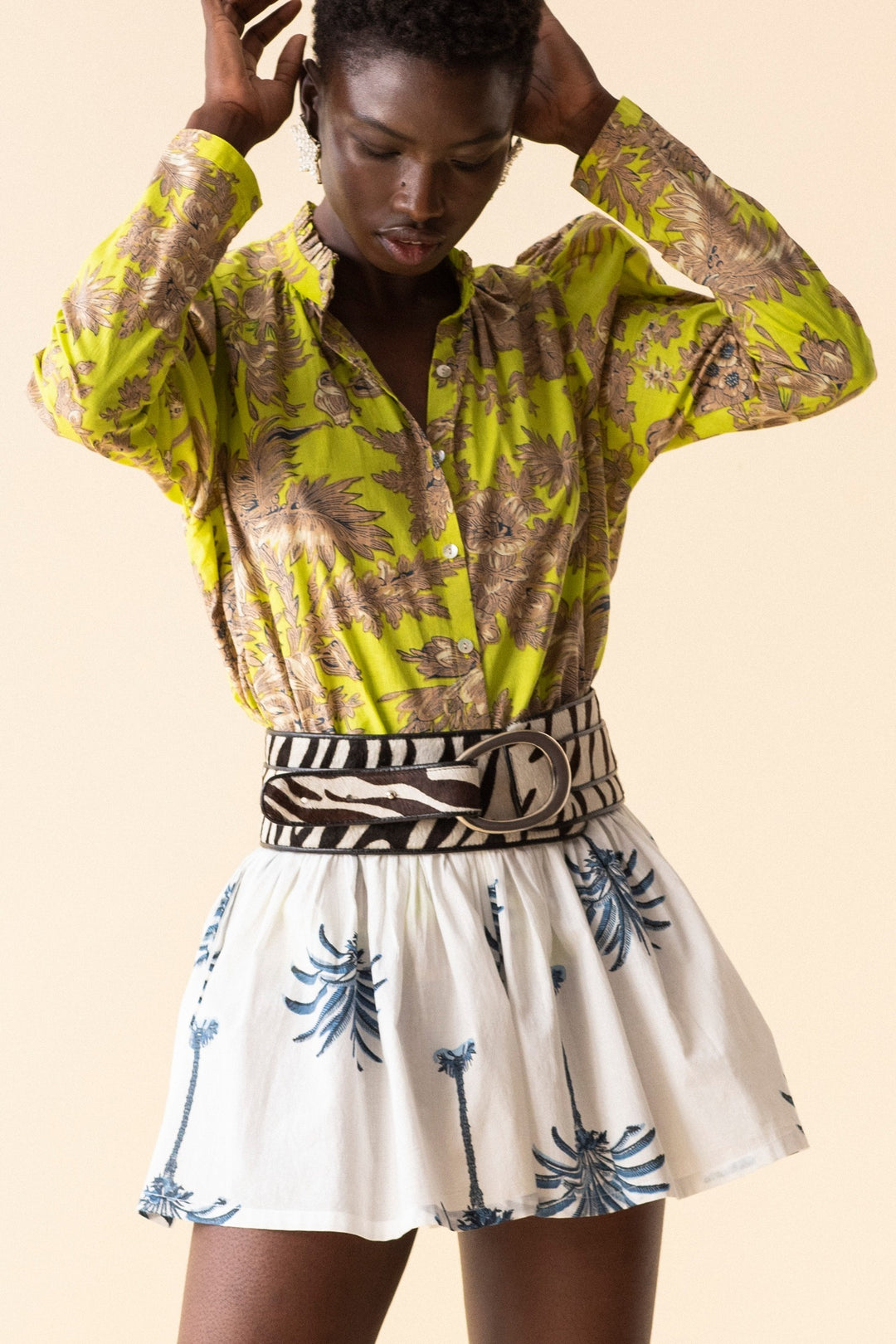 A person is wearing a vibrant yellow blouse adorned with a gold floral pattern, paired with the Christy Indigo Palm Short featuring blue palm tree designs. The shorts have a relaxed fit and come with a drawstring waistband. They accessorize with a wide patterned belt and have short hair, posing against a neutral background.