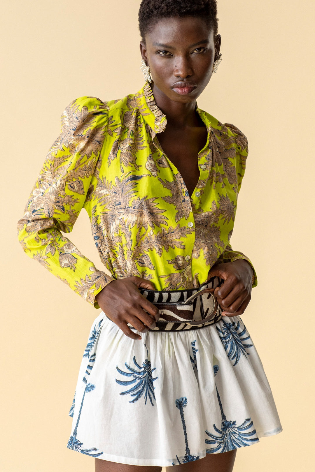 A person wearing an Annabel Acid Toile Shirt with a ruffled collar and a white skirt adorned with blue palm tree prints stands against a beige background. They accessorize with statement earrings and a zebra-patterned belt.