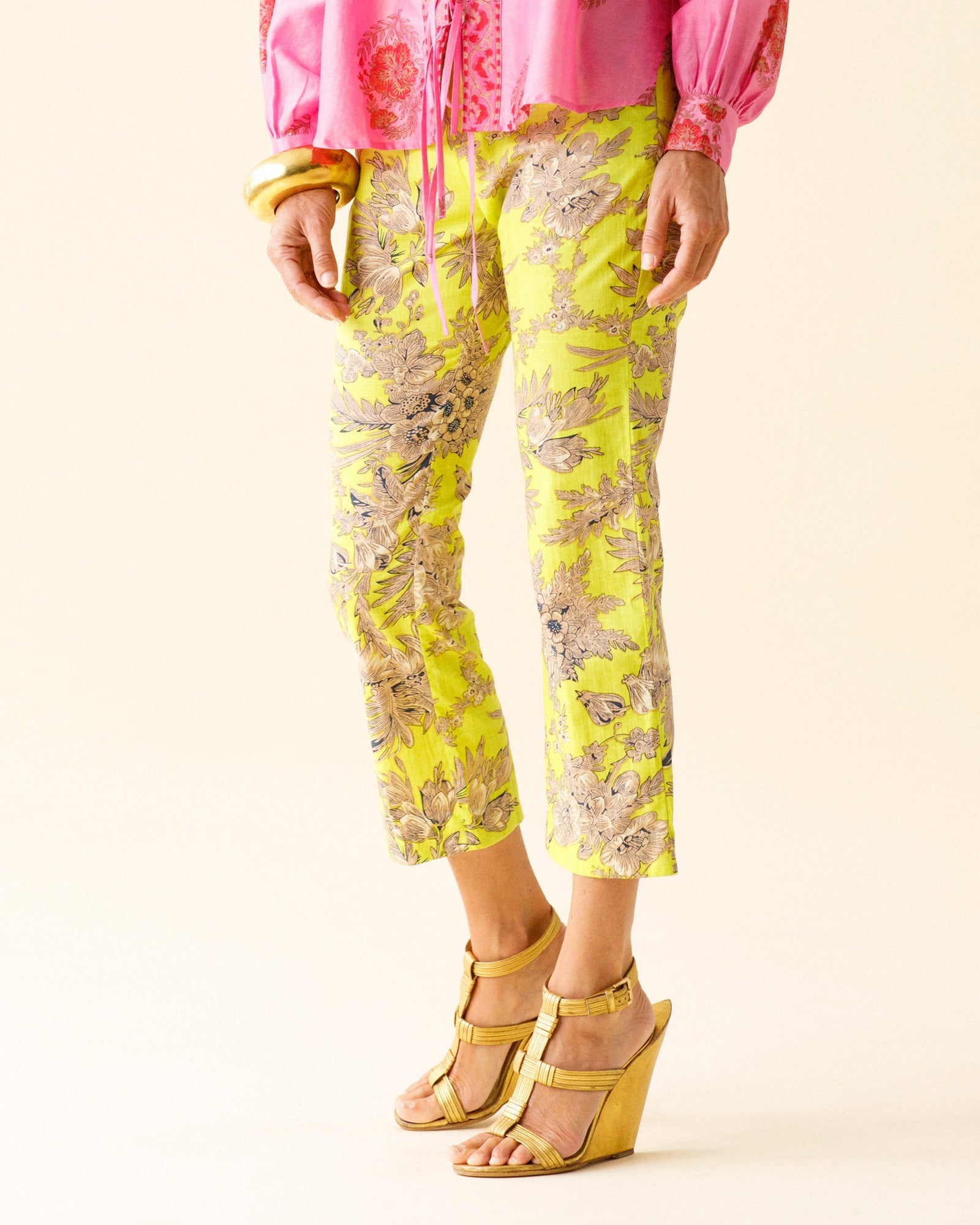 A person wearing Penny Acid Green Toile Pant made from hand-printed floral cotton with a slim cropped leg, a pink blouse, gold wedge sandals, and yellow bangles stands against a plain background.