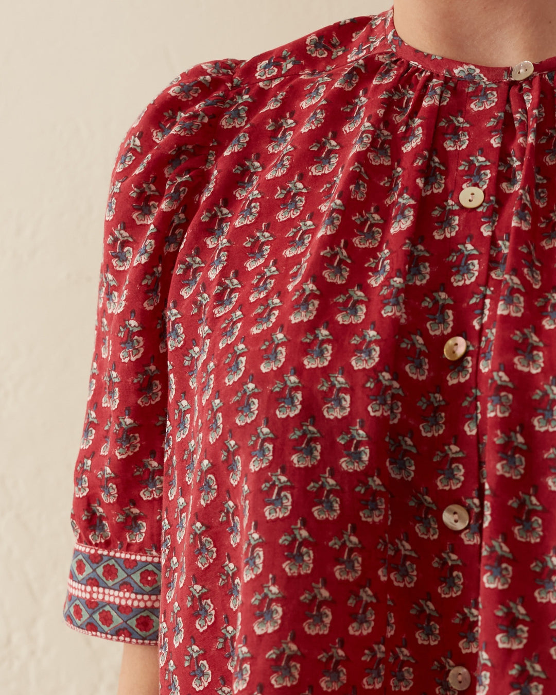 Close-up of the Winona Red Currant Shirt, featuring a hand-printed red floral design on cotton with a gathered neckline and elegant Mother of Pearl buttons. The puff sleeve showcases a blue geometric pattern at the cuff, set against a plain, light-colored wall backdrop.