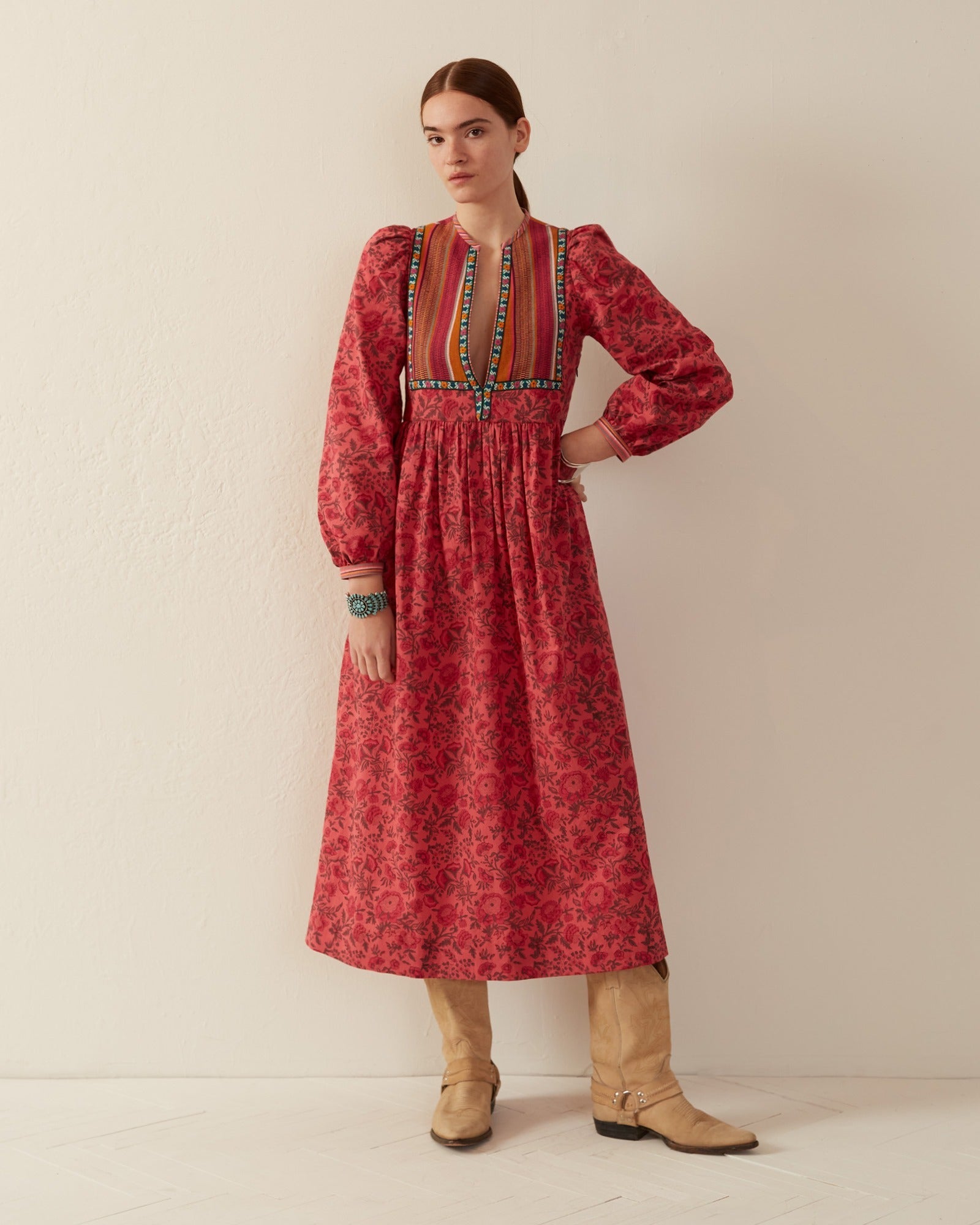 A person stands against a plain wall wearing the Winifred Raspberry Dress, a long, patterned red dress with colorful embroidery at the neckline. Crafted from hand-printed cotton and featuring a Guatemalan yoke, they maintain a neutral expression while wearing tan boots. Their arms rest by their side.