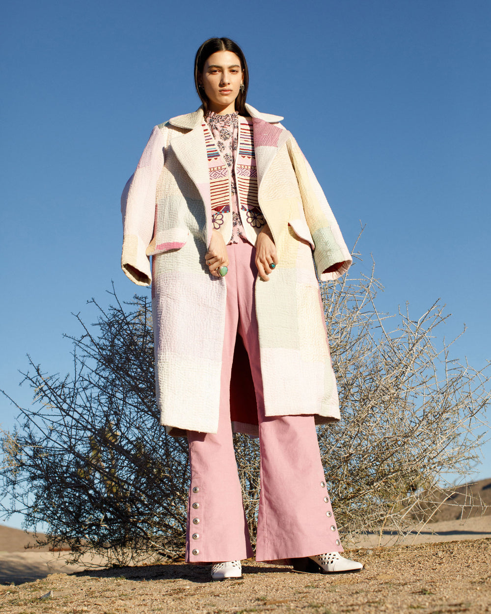 A person stands confidently in a desert landscape wearing the Officer Pastel Patchwork Kantha Coat over a patterned shirt and pink pants with buttons along the legs. They also have on white shoes, with a bush and clear blue sky in the background.