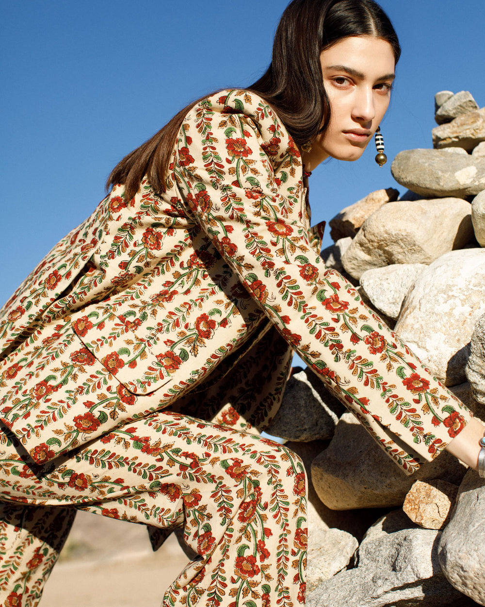 Wearing a Dree Climbing Ivy Blazer, which features hand-printed cotton with floral patterns and peak lapels, a person leans against a stack of rocks in the desert. The clear blue sky casts soft shadows as their long dark hair and single earring contribute to an effortlessly stylish look.