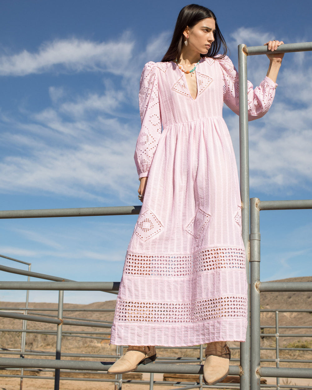 A woman stands outdoors on a metal railing, wearing the Winifred Blushed Eyelet Dress. She is gazing to the side against a backdrop of clear blue sky and barren landscape. Her tan boots perfectly complement the dress's intricate, handcrafted details.