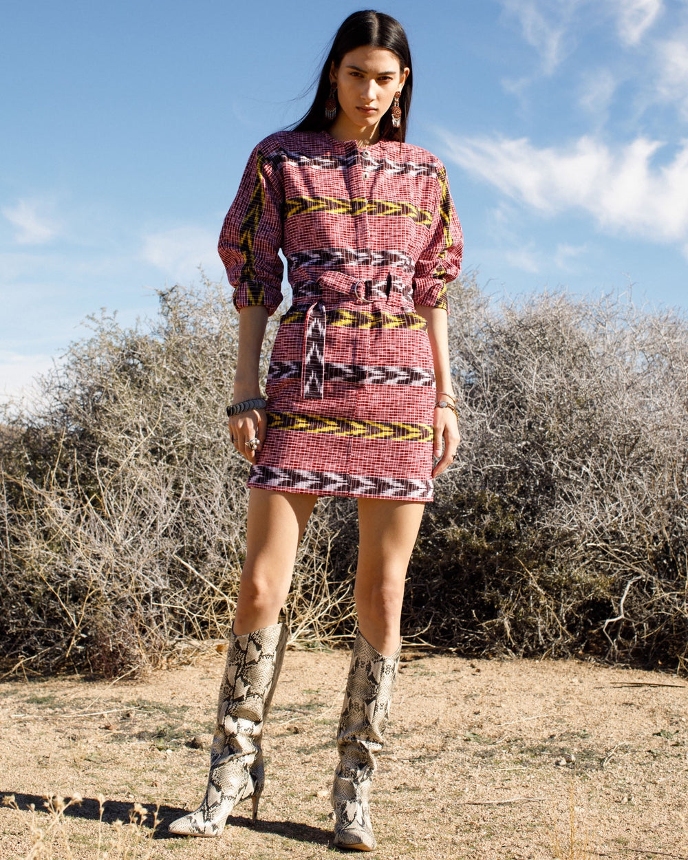 A person stands outdoors on a dry, grassy landscape, wearing the Laurent Pomegranate Ikat Dress with an adjustable D-ring belt and snake-patterned knee-high boots. The sky is blue with some clouds.