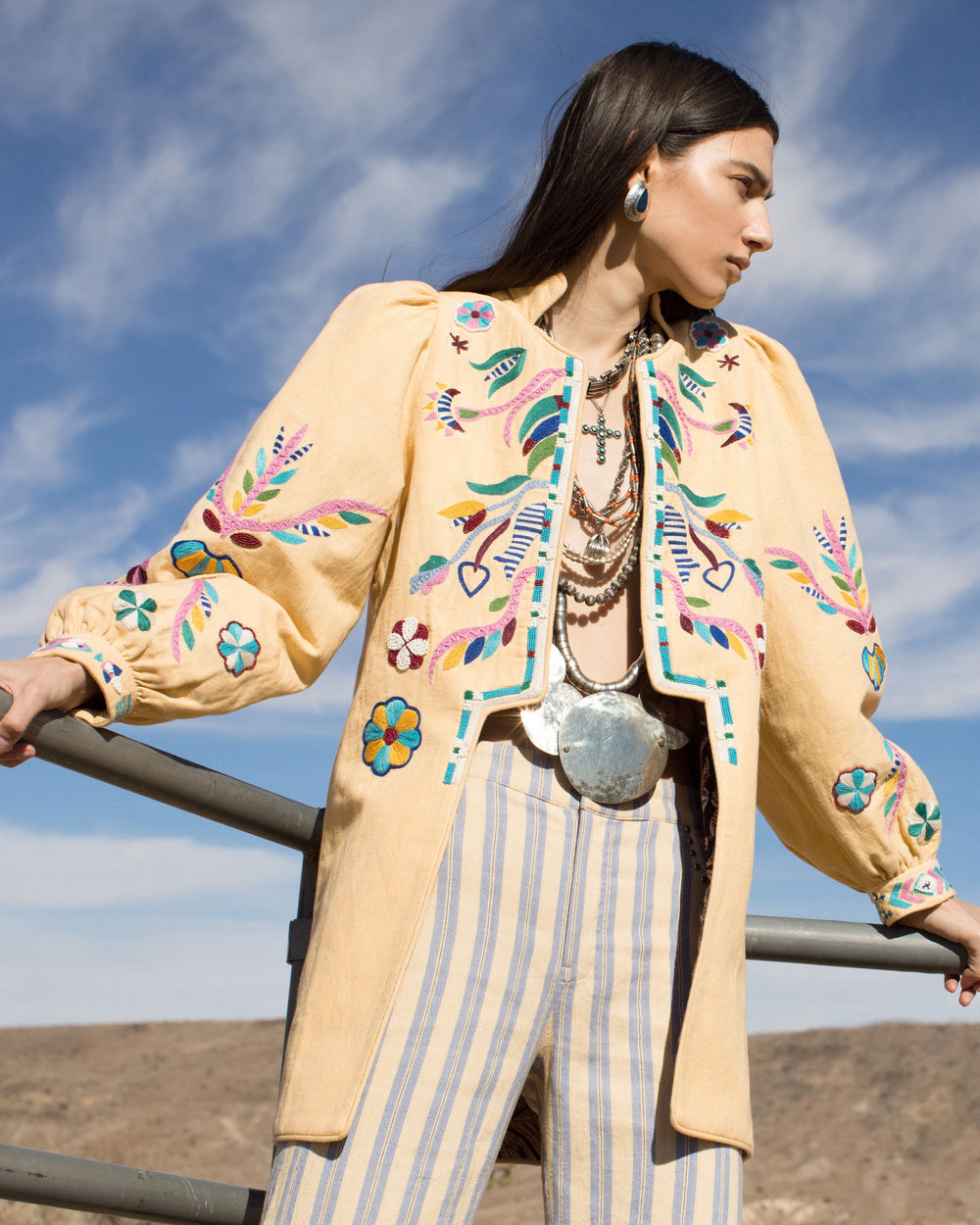 A woman stands outdoors wearing the Emmylou Butter Embroidered Coat, a colorful yellow jacket adorned with hand embroidery showcasing floral and geometric patterns. Her outfit is complemented by striped pants, layered necklaces, and puff sleeves. The background reveals a clear sky and rolling hills.