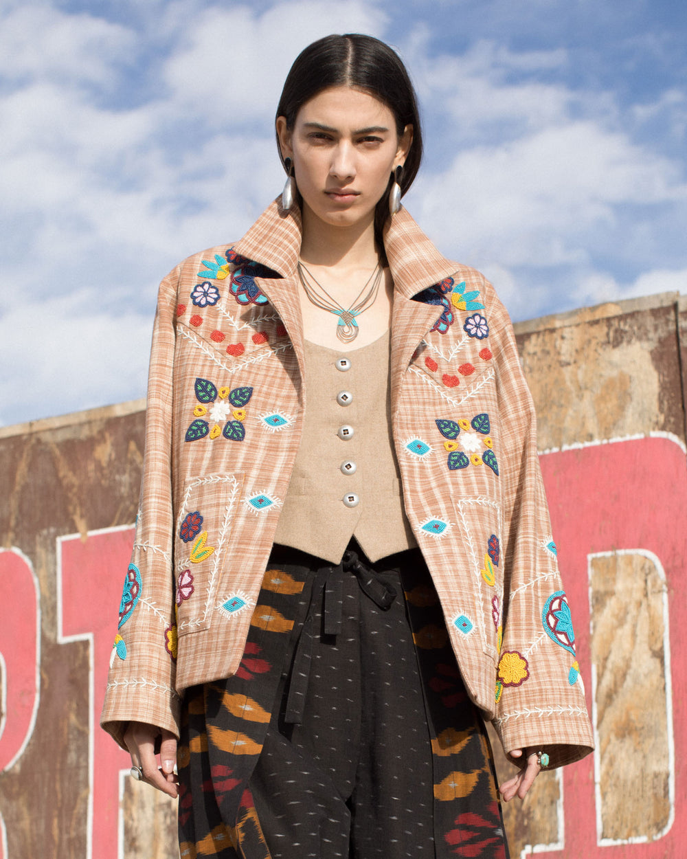 Amidst a rustic backdrop, a person stands confidently wearing the Western Tan Embroidered Jacket, which features colorful floral and geometric embroidery enhanced by delicate hand beading. This statement piece is layered over a beige top and paired with patterned trousers and layered necklaces, all under a partly cloudy sky.