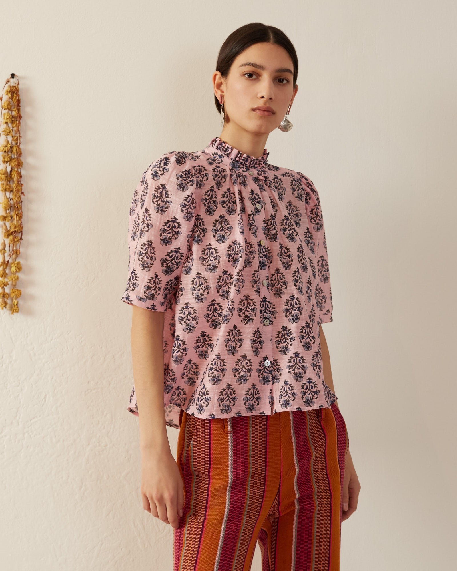 A person wearing the Winnie Pink Lily Shirt, made of hand-printed silk with ruffled collar details, along with striped pants, stands against a white wall. Dried flowers hang on the left side of the wall.