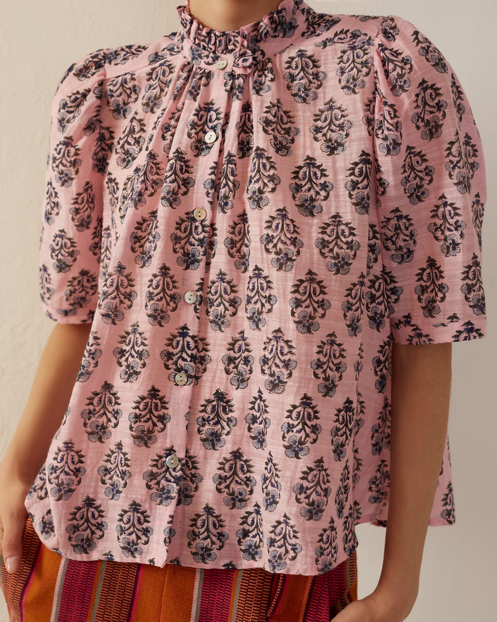 A person is wearing the Winnie Pink Lily Shirt, a pink hand-printed silk blouse featuring a ruffled collar and a striking black floral pattern. The elegant button-up front pairs beautifully with striped pants in vibrant orange and red hues.