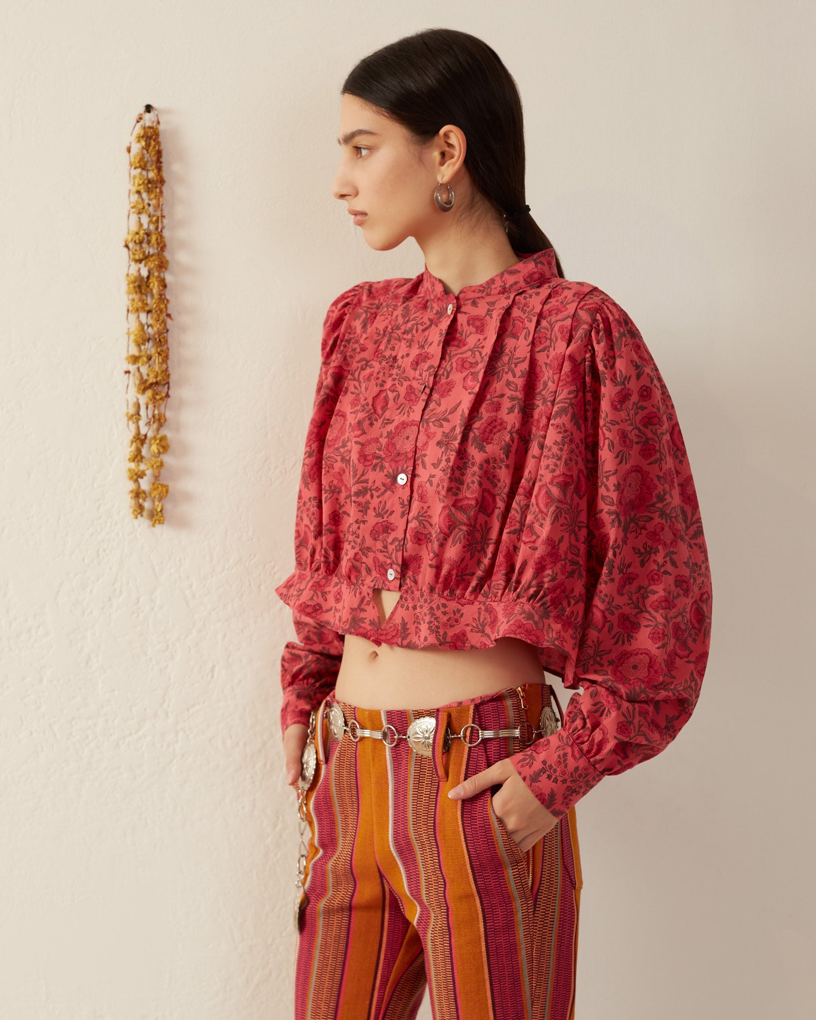 A person with long dark hair stands sideways, wearing the Rhiannon Raspberry Blouse, which features pleating details and is paired with striped pants in orange and maroon tones. They are accessorized with a silver belt and earrings, while a garland hangs on the white wall, enhancing the Victorian silhouette charm of the scene.