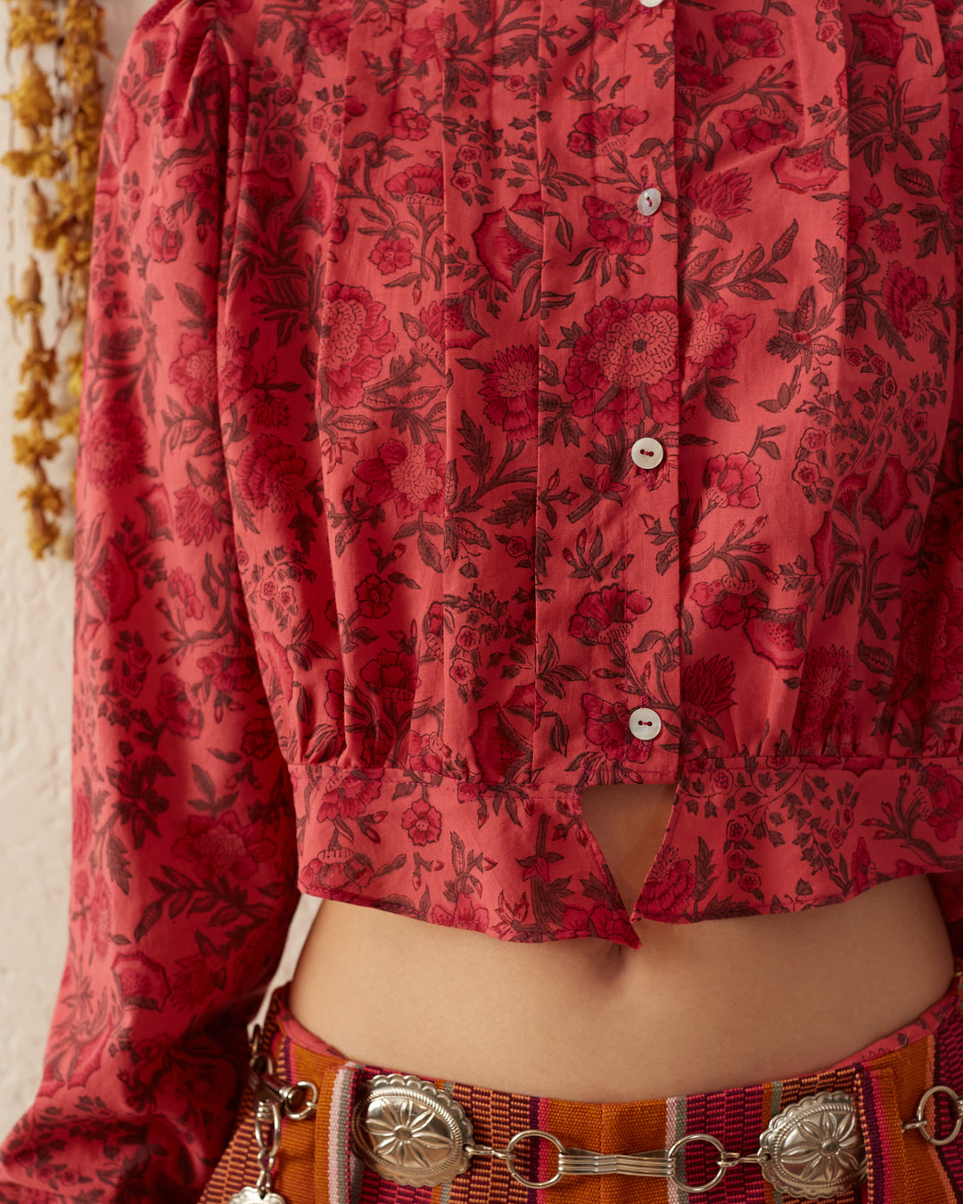 The Rhiannon Raspberry Blouse is a red patterned masterpiece adorned with floral designs. Its partial button-up style highlights the Victorian silhouette, and when paired with a belt featuring decorative metal elements, this hand-printed cotton blouse exudes timeless elegance.