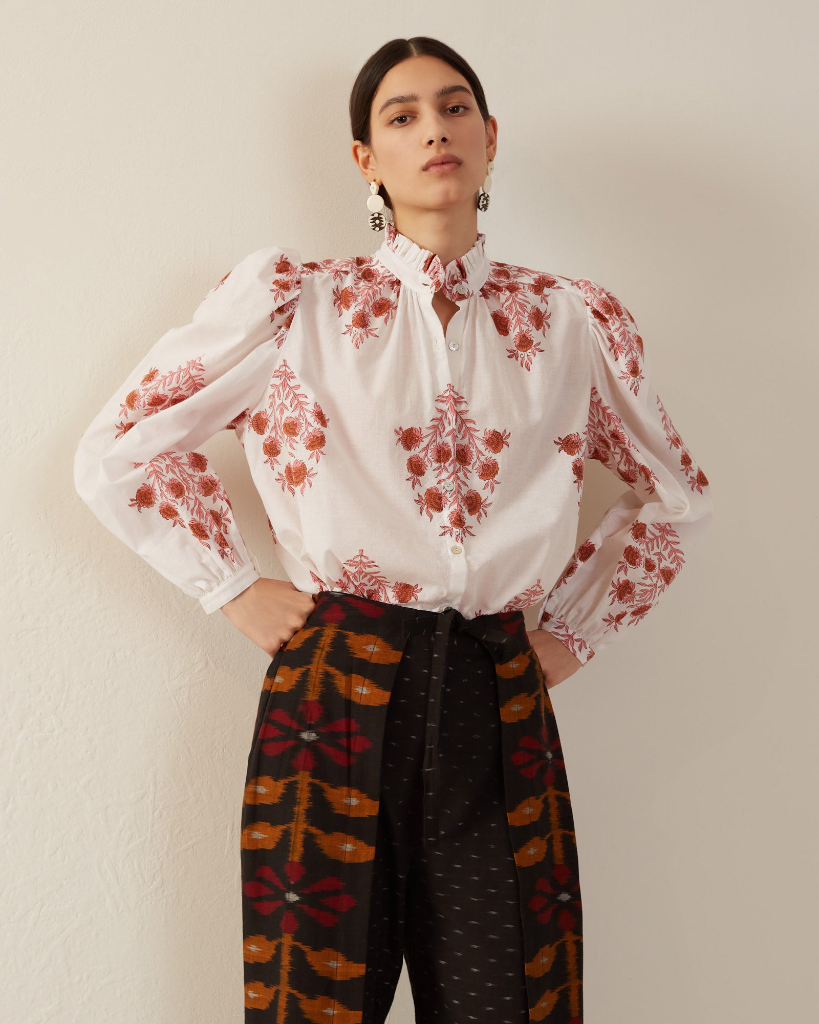 A person wearing the Annabel Pink Dahlia Blouse, adorned with red floral patterns and paired with high-waisted pants made from hand-printed cotton, poses confidently against a plain, light-colored background. Their long, dark hair complements their statement earrings, and the blouse features a delicate Mother of Pearl button.