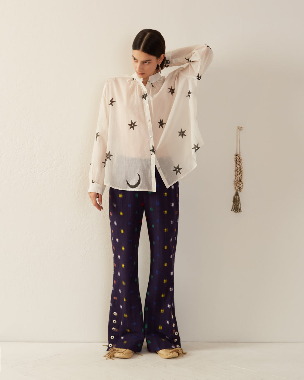 In front of a white wall, someone is dressed in the Kiki Stargazer Blouse, featuring sheer star patterns, paired with dark patterned pants. A decorative tassel hangs in the background while Mother of Pearl buttons enhance the outfit’s elegance.