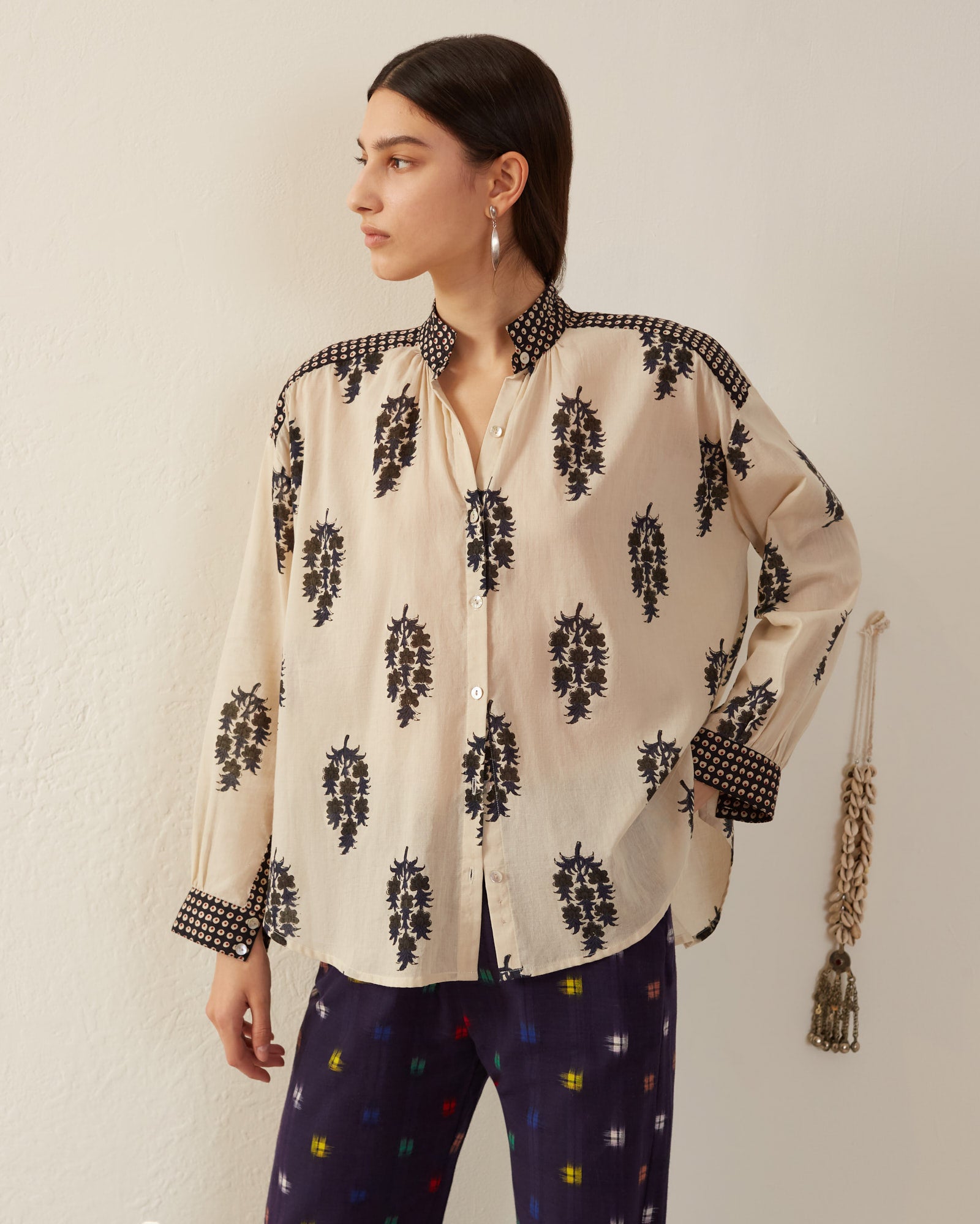 A person wearing the Kiki Black Dahlia Shirt, featuring a cream color with black floral patterns and checkered designs on the shoulders and cuffs, highlights Mother of Pearl buttons. Paired with dark pants showcasing a vibrant pattern, they pose against a neutral backdrop with a rope accent on the wall.