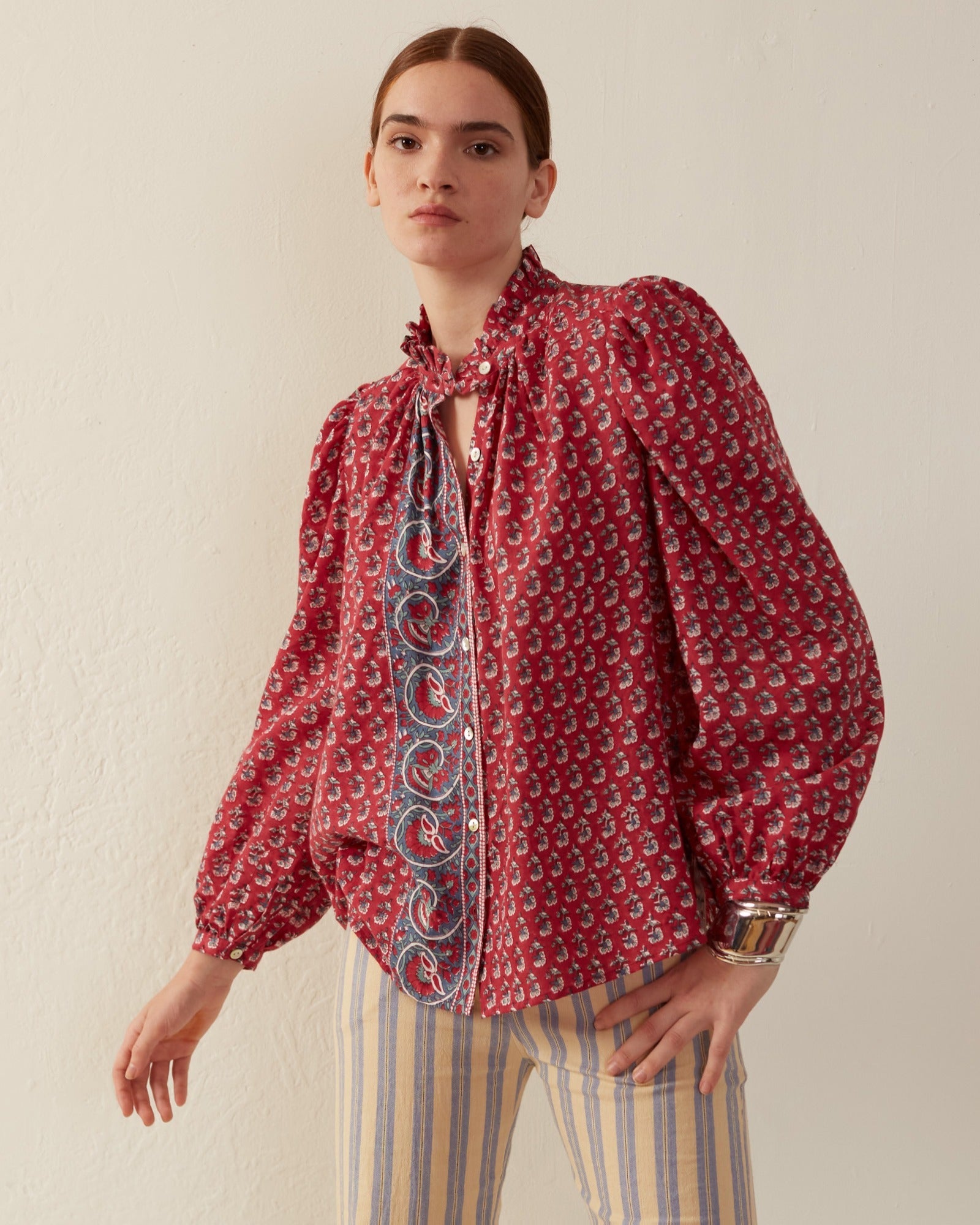 A person stands against a plain background, wearing the Annabel Red Currant Shirt, which features puffed sleeves and a ruffled collar, paired with light-colored striped pants. They have long hair pulled back and wear a metallic bracelet.