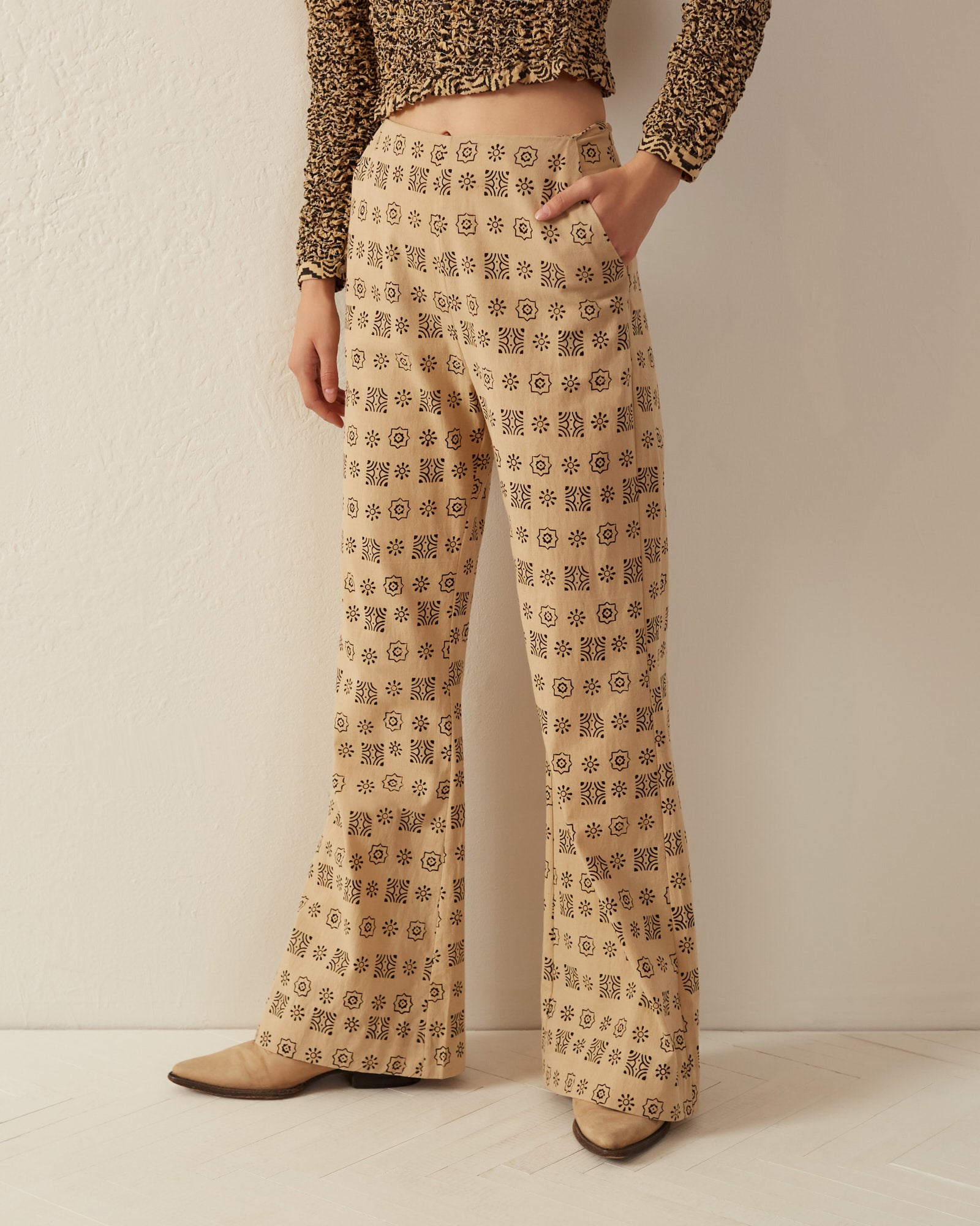 A person is wearing the Charlie Jute Bandana Pant, high-waisted beige trousers featuring a black geometric pattern. These flared-leg pants, made from hand-printed cotton and complete with side pockets, are paired with a textured brown top as they stand against a pale background with one hand in their pocket.