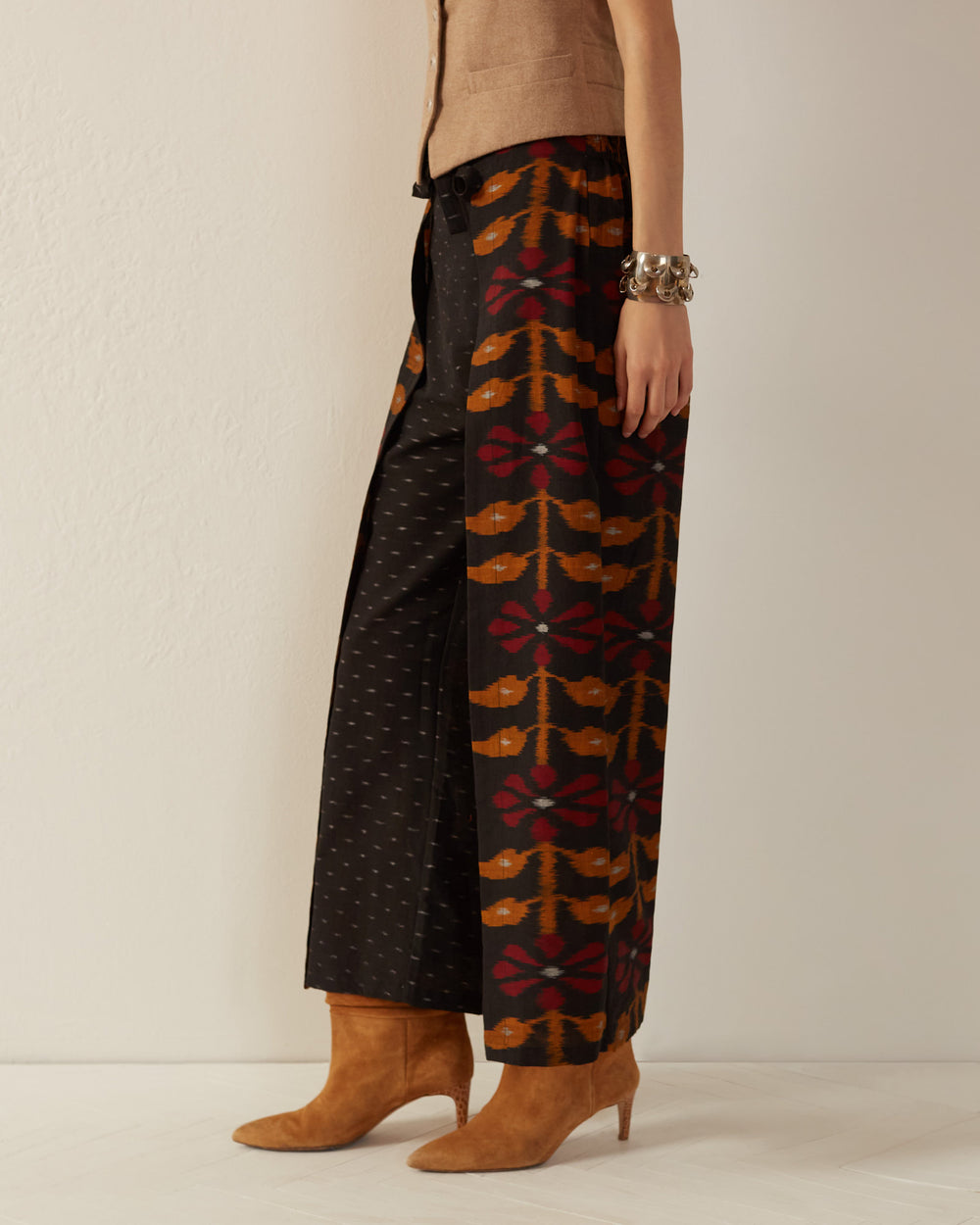 A person is wearing a brown vest and the Bonnie Black Bloom Wrap Pants, which feature a colorful, geometric pattern reminiscent of Indian ikat. They pair it with brown heeled boots and a chunky bracelet, set against a plain, off-white wall.