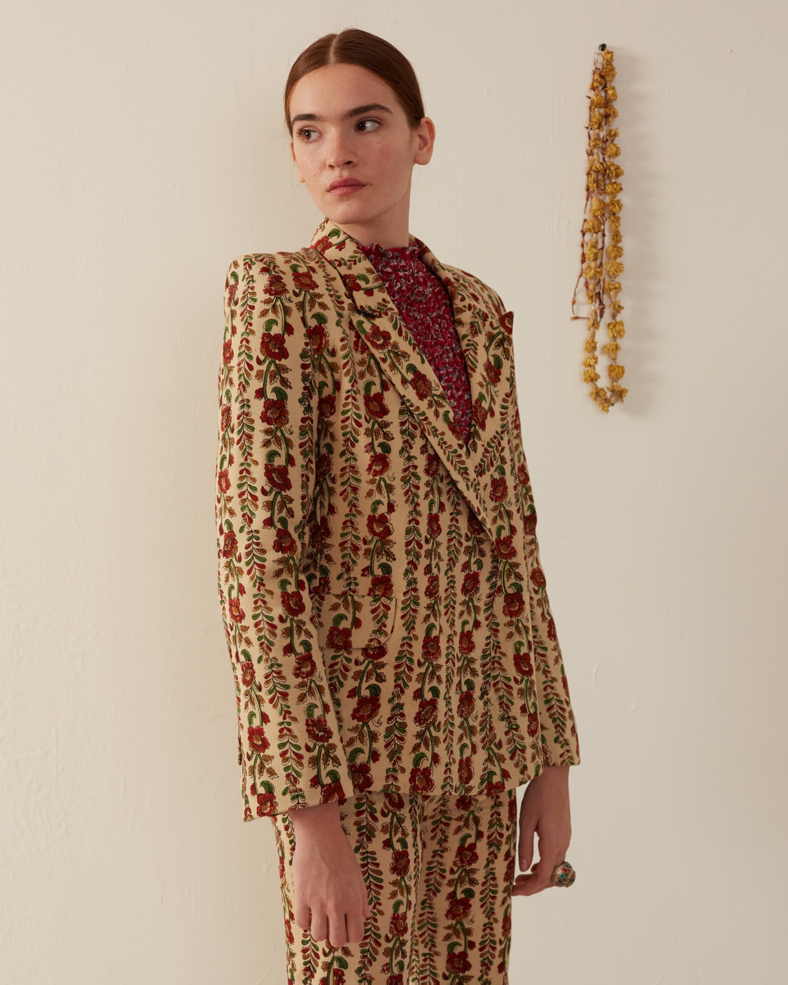 A person stands against a light-colored wall, wearing a Dree Climbing Ivy Blazer in hand-printed cotton with floral designs in red and green. The suit's peak lapel adds a classic touch as they gaze to the side. A string of dried flowers hangs on the wall behind them.