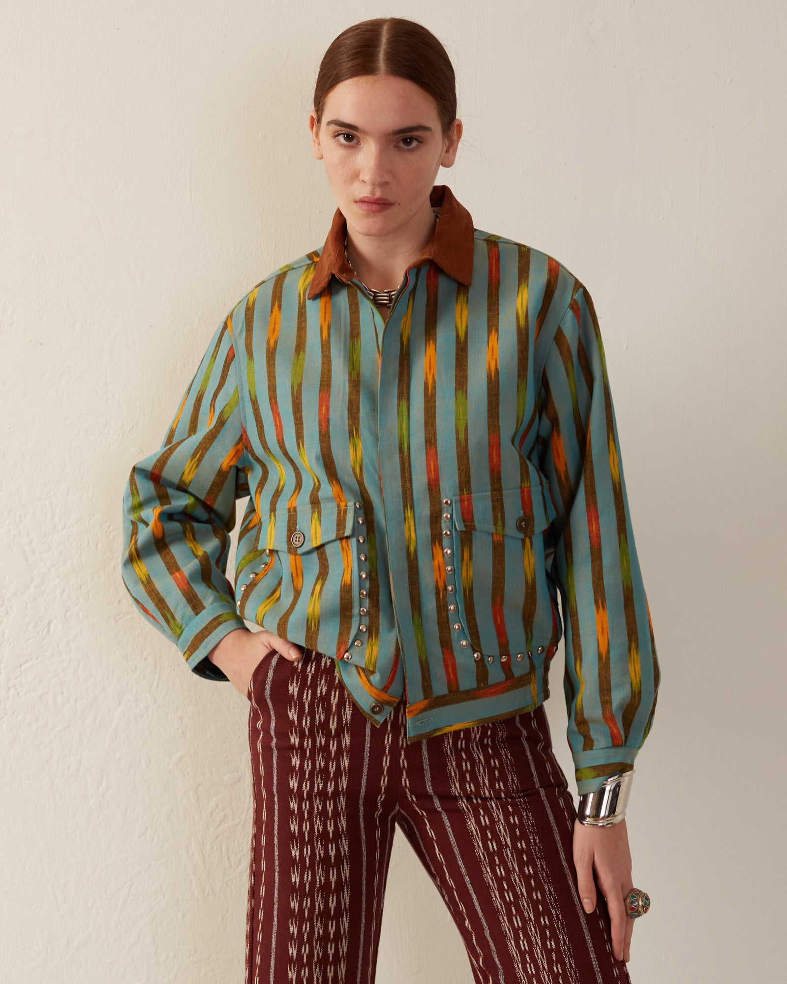 A person is standing against a white wall, wearing the Lucky Aqua Ikat Jacket featuring multicolored stripes and a brown collar. They pair it with patterned maroon pants that have a gathered elastic waist. With one hand on their hip, they confidently gaze at the camera.