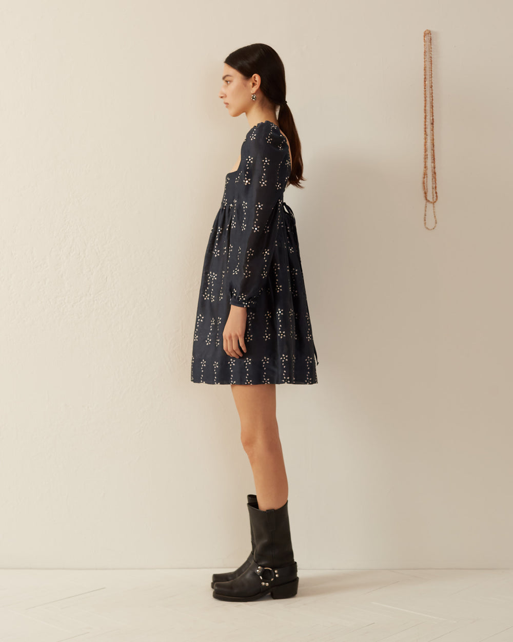 A woman in profile stands against an off-white wall, wearing the Christina Black Bloom Dress, which features a dark blue pattern crafted with the hand printing process. She pairs the dress with black boots. Her hair is tied back, and a long beaded necklace hangs on the wall beside her.