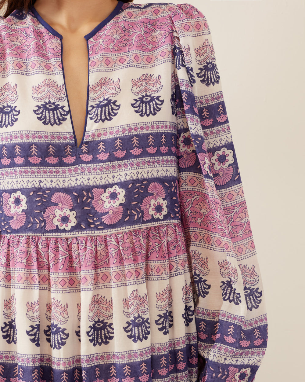 A close-up of a person wearing the Winifred Lavender Dawn Dress, featuring a vibrant V-neckline crafted from hand-printed cotton. The dress showcases intricate floral motifs and geometric shapes in pink, purple, and white, with long sleeves gathered at the cuffs for an elegant finish.