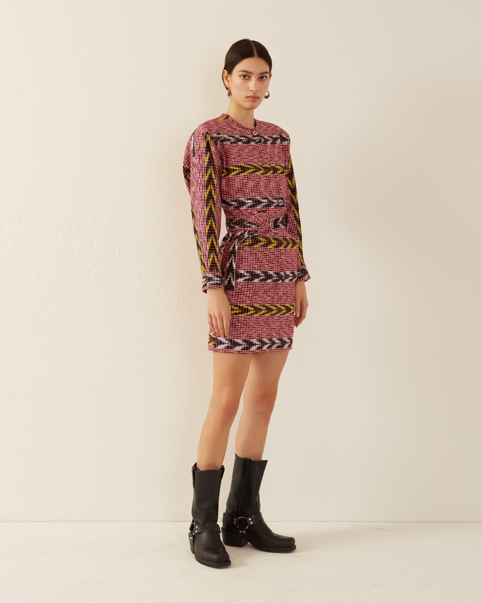 A person stands against a plain backdrop wearing the Laurent Pomegranate Ikat Dress, featuring a pink and yellow pattern made from hand-woven Guatemalan ikat, paired with black boots. They have dark hair pulled back and are looking towards the camera.