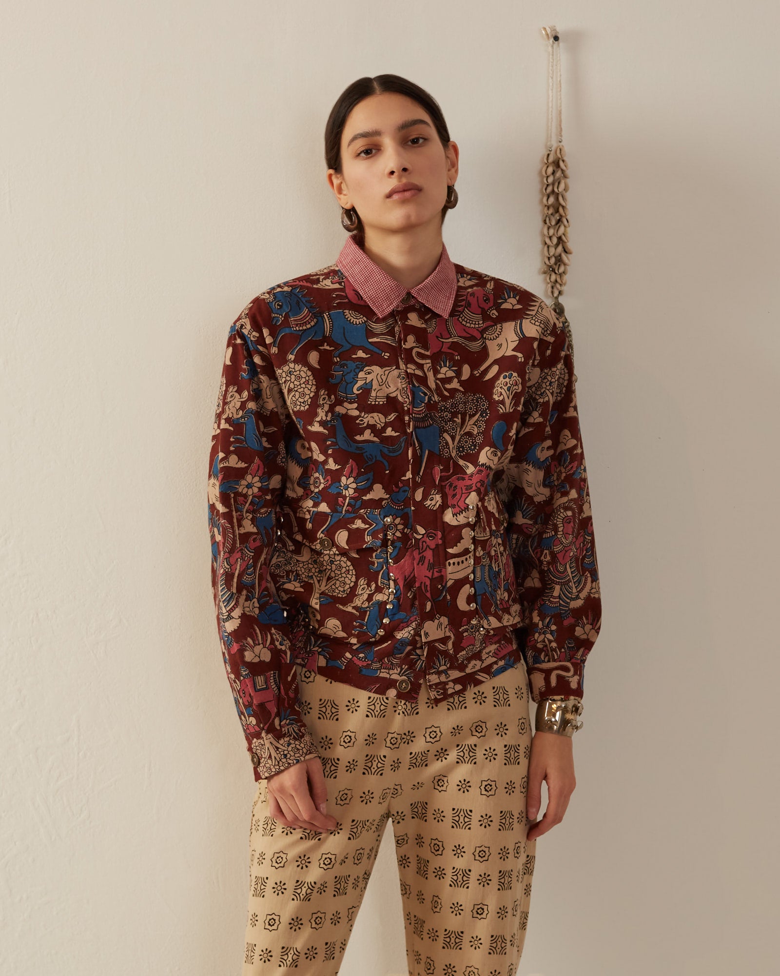 A person with long hair stands against a plain wall, wearing their Lucky Coffee Kalamkari Jacket adorned with floral and animal designs. The jacket, crafted using traditional methods on hand-printed cotton, complements a pink collared shirt and beige pants featuring geometric prints. A string of shells or beads hangs on the wall.