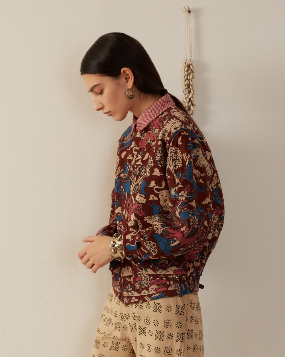 A person with long dark hair, wearing a Lucky Coffee Kalamkari Jacket made of hand-printed cotton in maroon and blue over a red checked shirt, stands sideways against a cream-colored wall. They have large hoop earrings and beige pants with black designs. A decorative hanging is in the background.