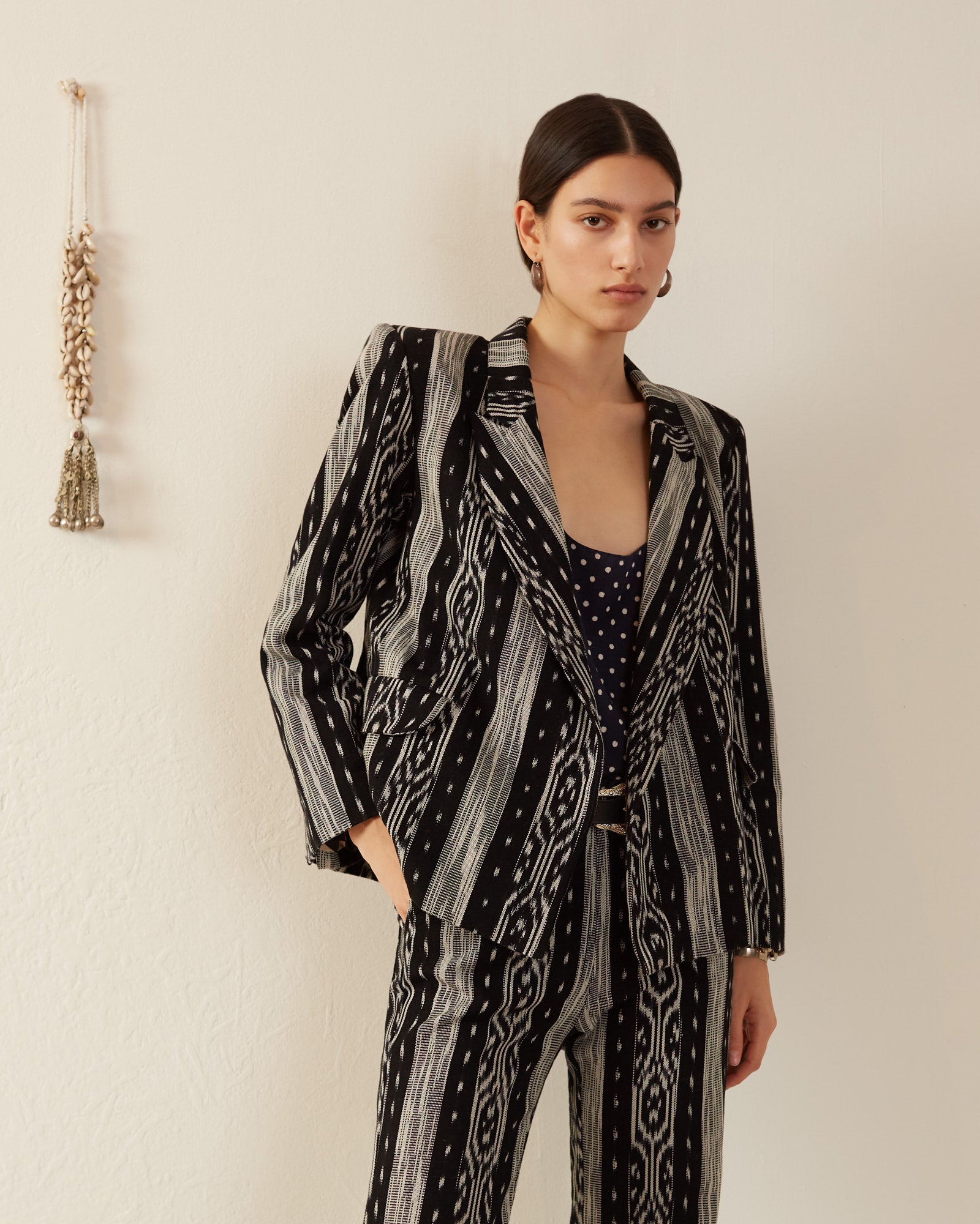 A person with long dark hair stands against a beige wall, wearing a stylish Dree Obsidian Ikat Blazer over a polka dot top. A decorative tassel, reminiscent of Guatemalan ikat designs, hangs on the wall to their left.