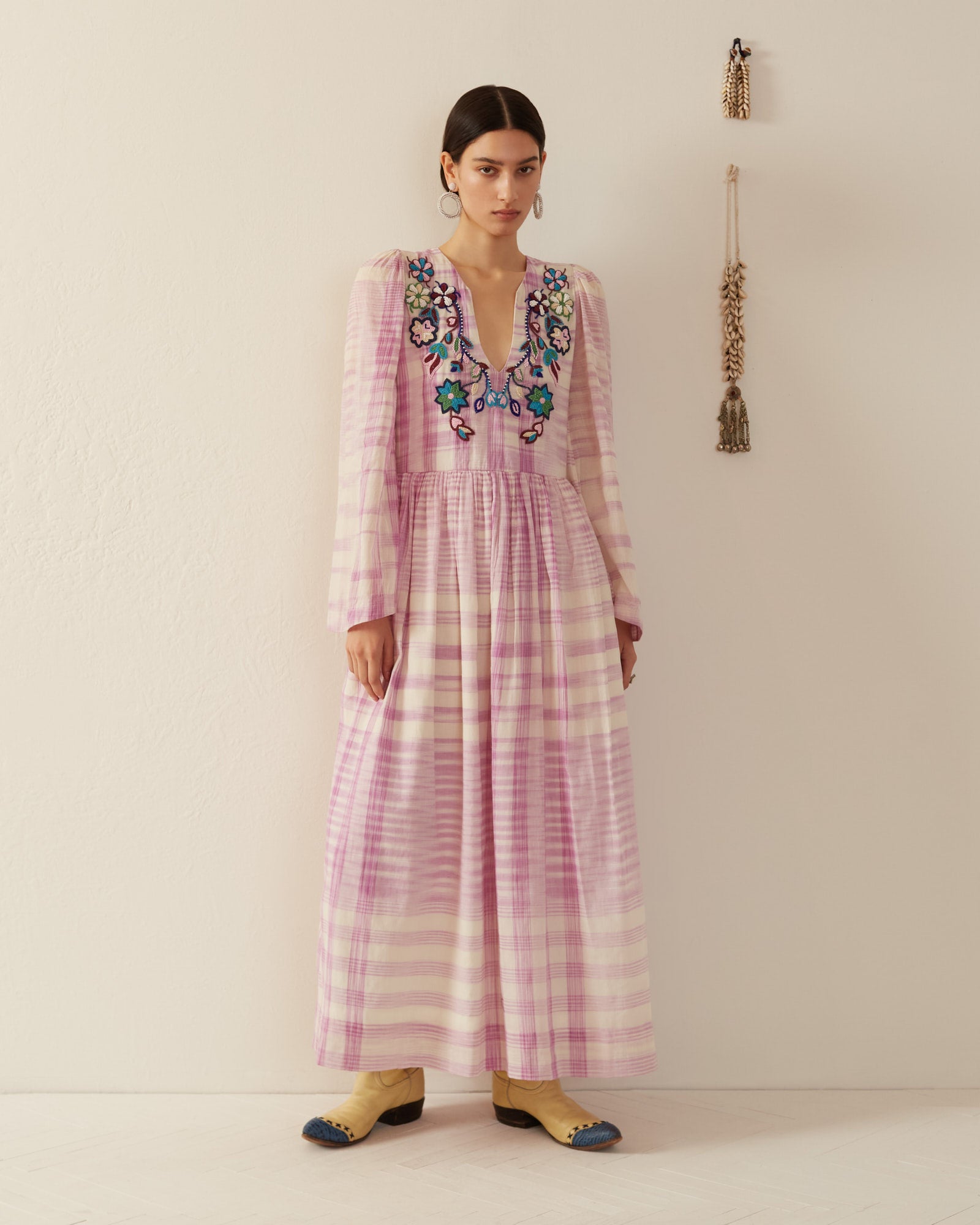 A person wearing the Savannah Purple Prairie Dress, featuring a long silhouette with floral embroidery and bell sleeves, stands against a beige wall. They have on brown boots, and there is a small wall decoration beside them.