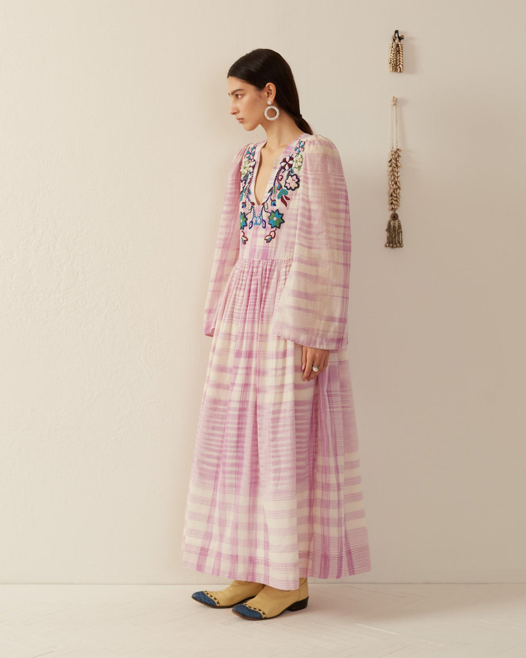 A woman stands against a cream wall wearing the Savannah Purple Prairie Dress, a flowing pink and white plaid garment adorned with floral embroidery and hand beading. Her arms are at her sides, framed by bell sleeves. She accessorizes with large hoop earrings and brown shoes, while tassels hang on the wall to complete the scene.