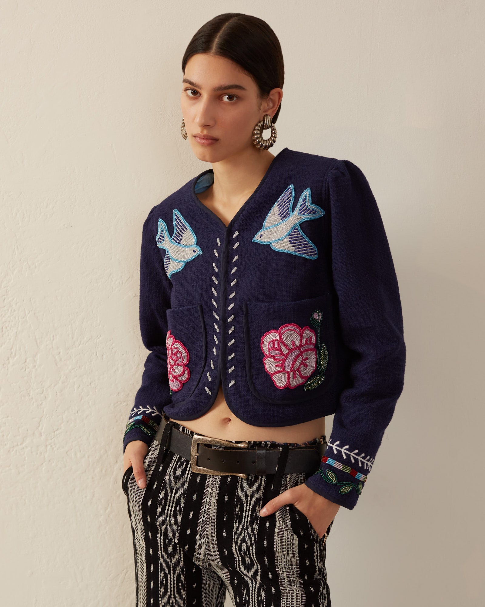 Against a plain background, a person poses wearing the Stella Swallow Rose Jacket, a navy blue piece adorned with hand-embroidered birds and flowers. They complement it with striped, woven cotton pants and large earrings while keeping their hands in their pockets as they gaze at the camera.