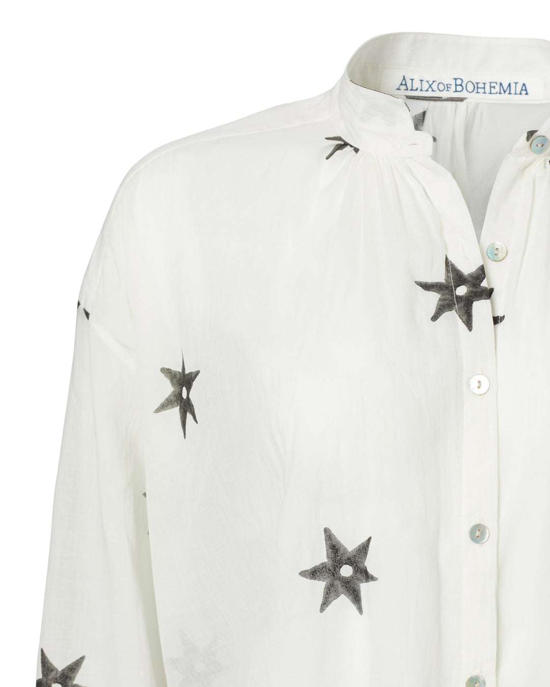 The Kiki Stargazer Blouse showcases a partial view of white fabric adorned with black star patterns and displays the "ALIX of BOHEMIA" brand label at the neckline. Skillfully crafted using traditional techniques from hand-printed cotton, it is adorned with elegant Mother of Pearl buttons that add a touch of timeless charm.