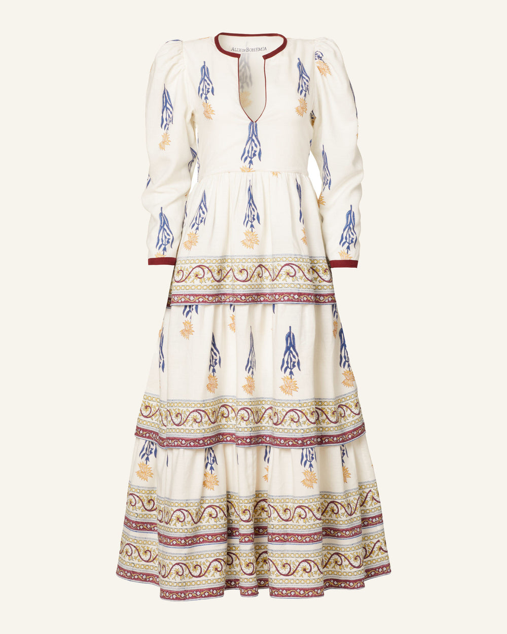 The Tallulah Falling Daffodil Dress is a long, white floral dress featuring blue patterns and detailed red, yellow, and blue designs along the V-neckline and sleeves. It includes long puff sleeves and a tiered ruffle skirt made from hand-printed Tencel.