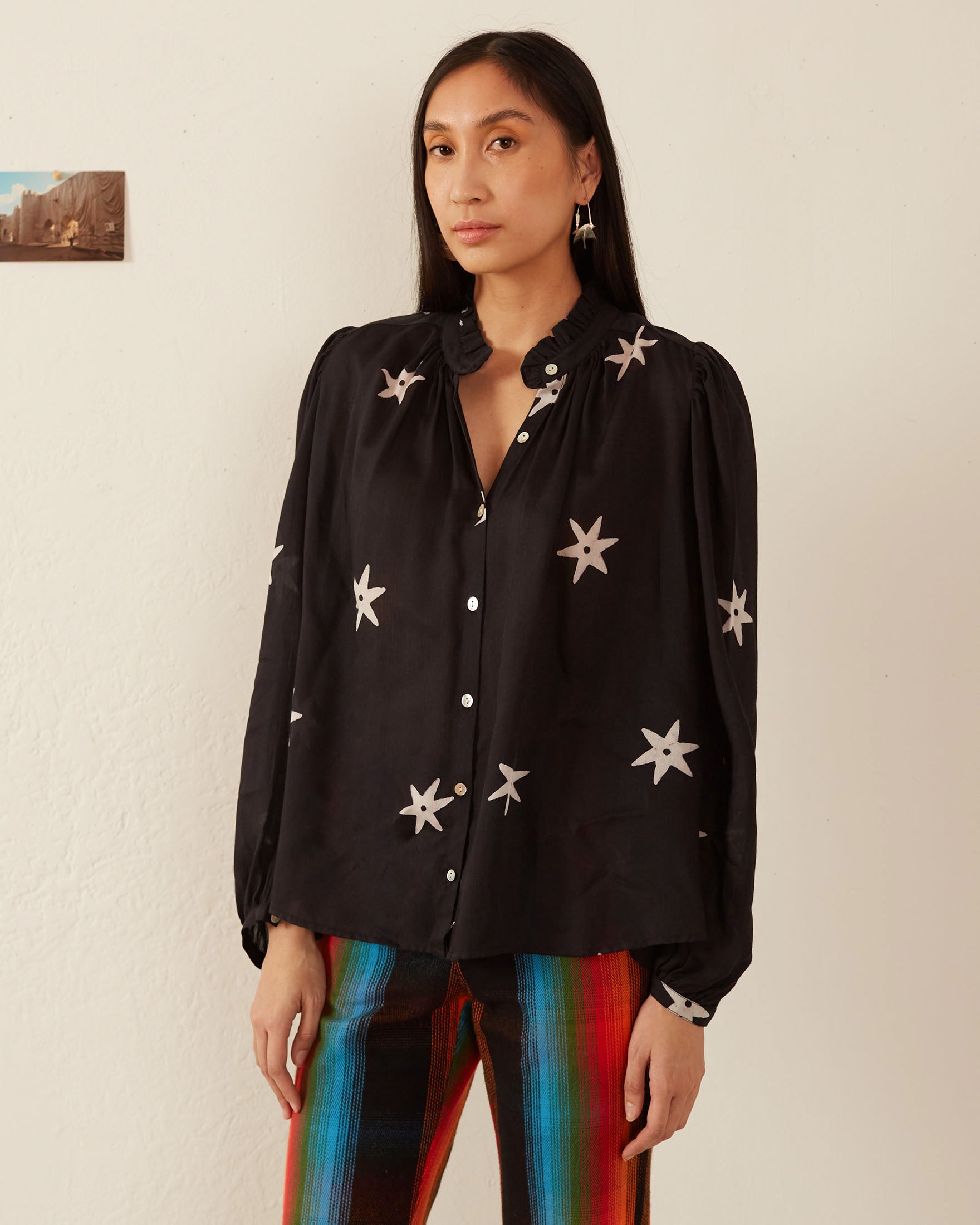 A person with long hair wears the Annabel Night Star Shirt, a black blouse adorned with star patterns and puffed sleeves, paired with colorful striped pants. They stand against a light-colored wall with a small picture hanging on it.