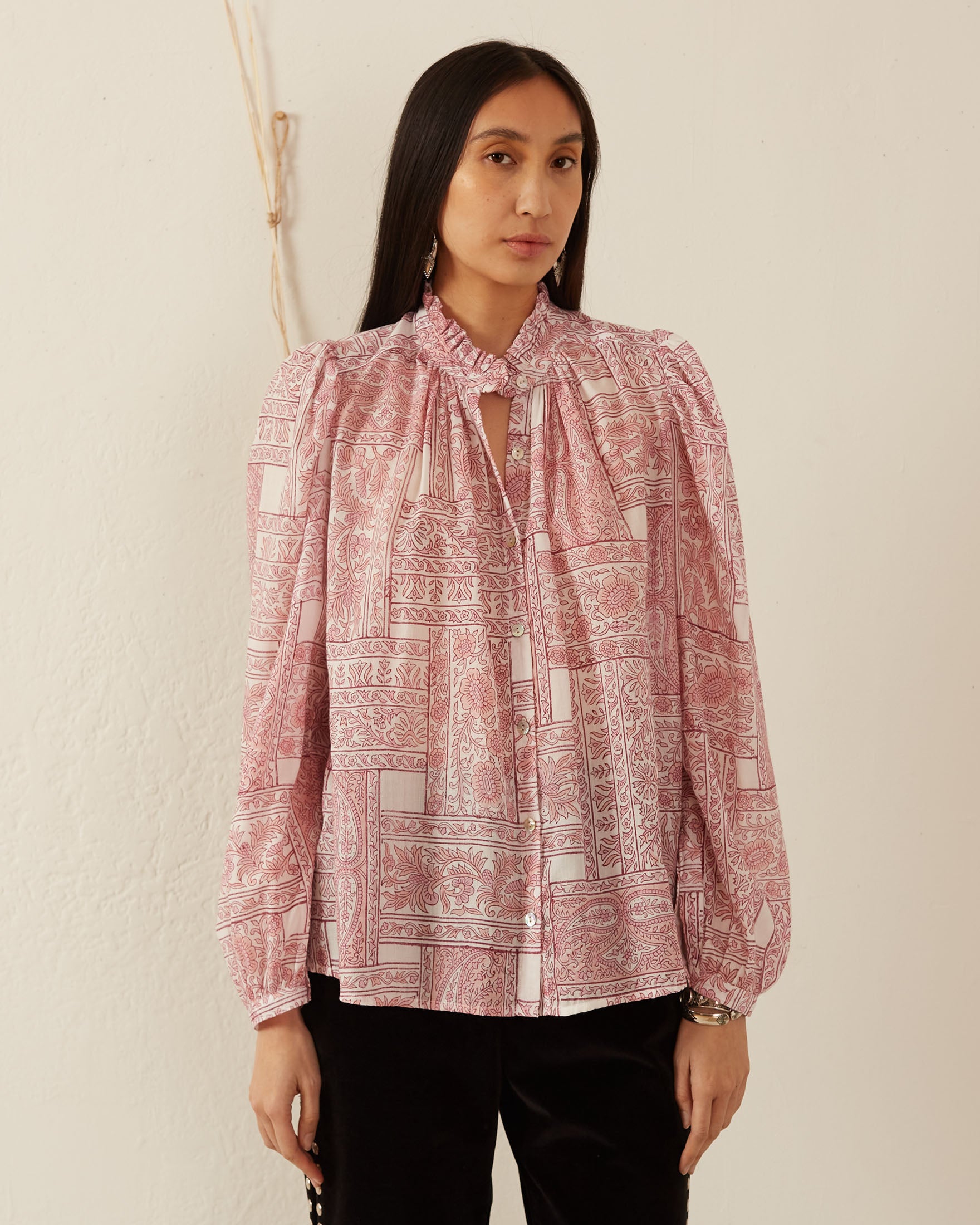 A person with long, dark hair stands against a light-colored wall, wearing the Annabel Pink Crossroad Shirt—a hand-printed Tencel blouse featuring a ruffled collar and puffed sleeves—paired with dark pants.