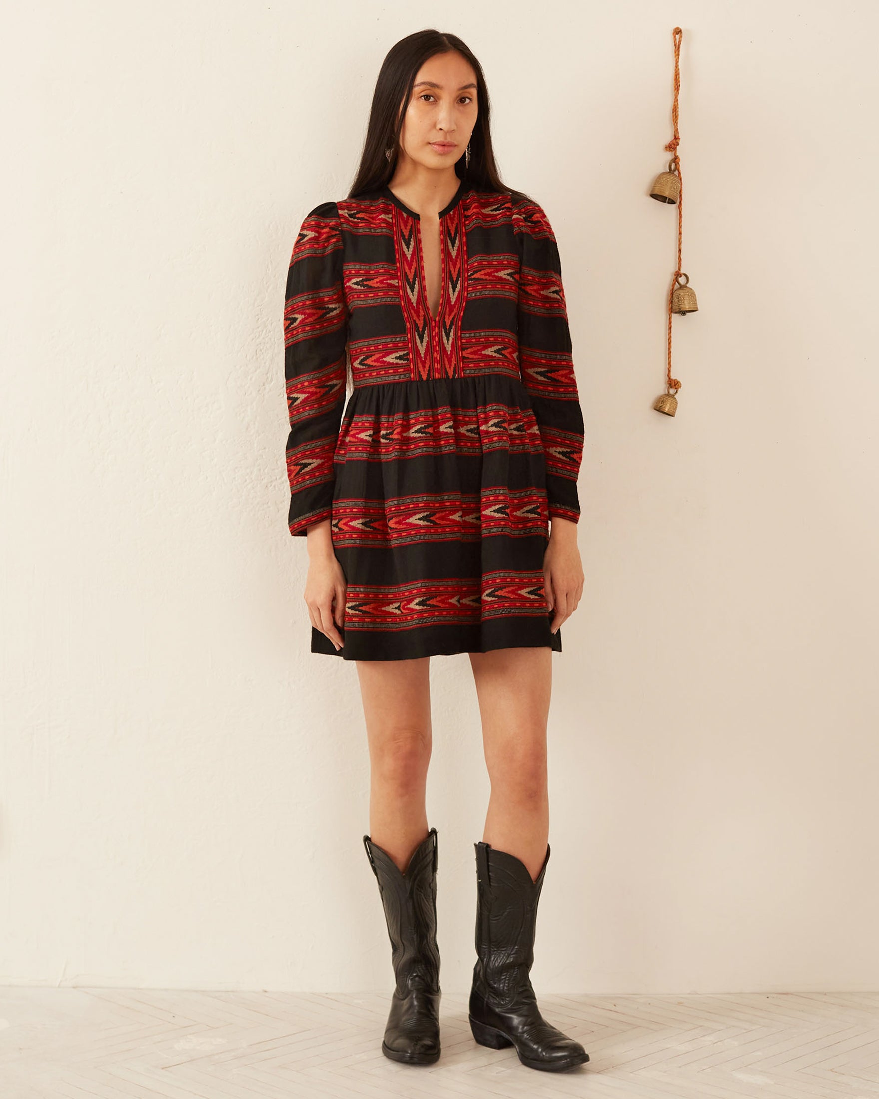 A person stands against a cream-colored wall, wearing a red and black patterned Winslow Arrow Dress with long sleeves, crafted using traditional methods. Black cowboy boots complete the look. A small wall hanging with bells is visible to the right.