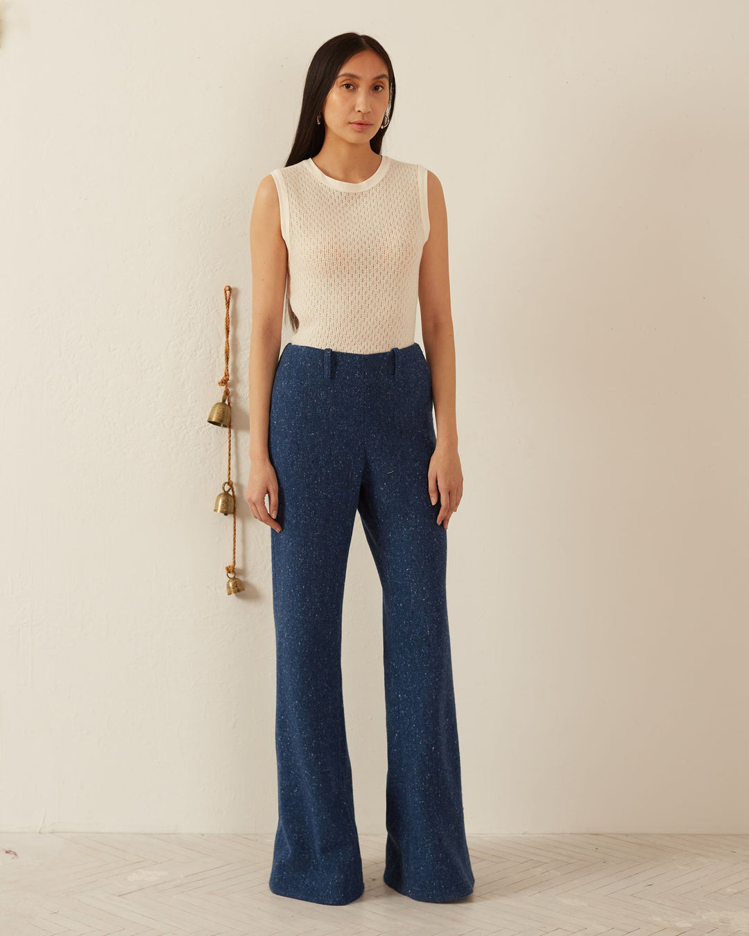 A person with long dark hair stands against a light wall, wearing a sleeveless cream top and the Charlie Blueberry Tweed Pant featuring a subtle side zipper. Bells hang on the wall beside them, and the wooden floor adds warmth to the minimalist setting.