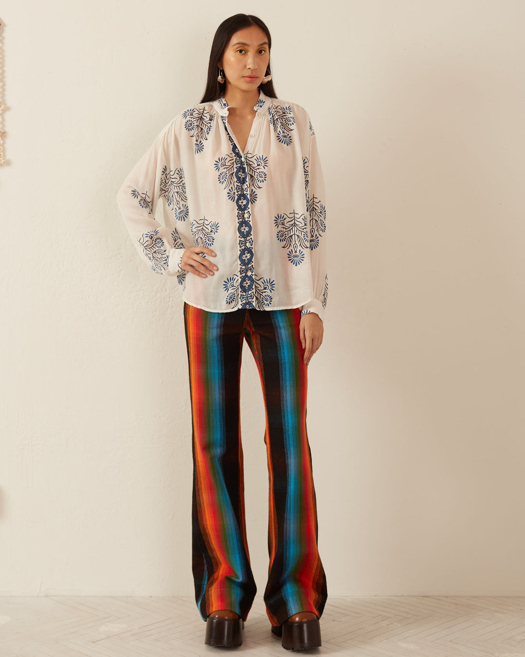 A woman stands against a plain wall, wearing the Kiki Black and Blue Shirt, featuring delicate Mother of Pearl buttons and a blue floral design. Her vibrant, multicolored striped pants complement her dark platform shoes and long earrings. Her straight hair frames her face as she looks confidently at the camera.