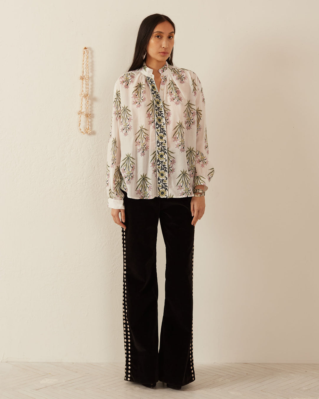 A woman stands against a beige wall with a beaded decoration, wearing the floral-patterned, long-sleeved Kiki Winter Lily Shirt adorned with Mother of Pearl buttons. She's paired it with black pants featuring decorative beadwork along the sides. The floor boasts a light-colored, subtle geometric pattern.