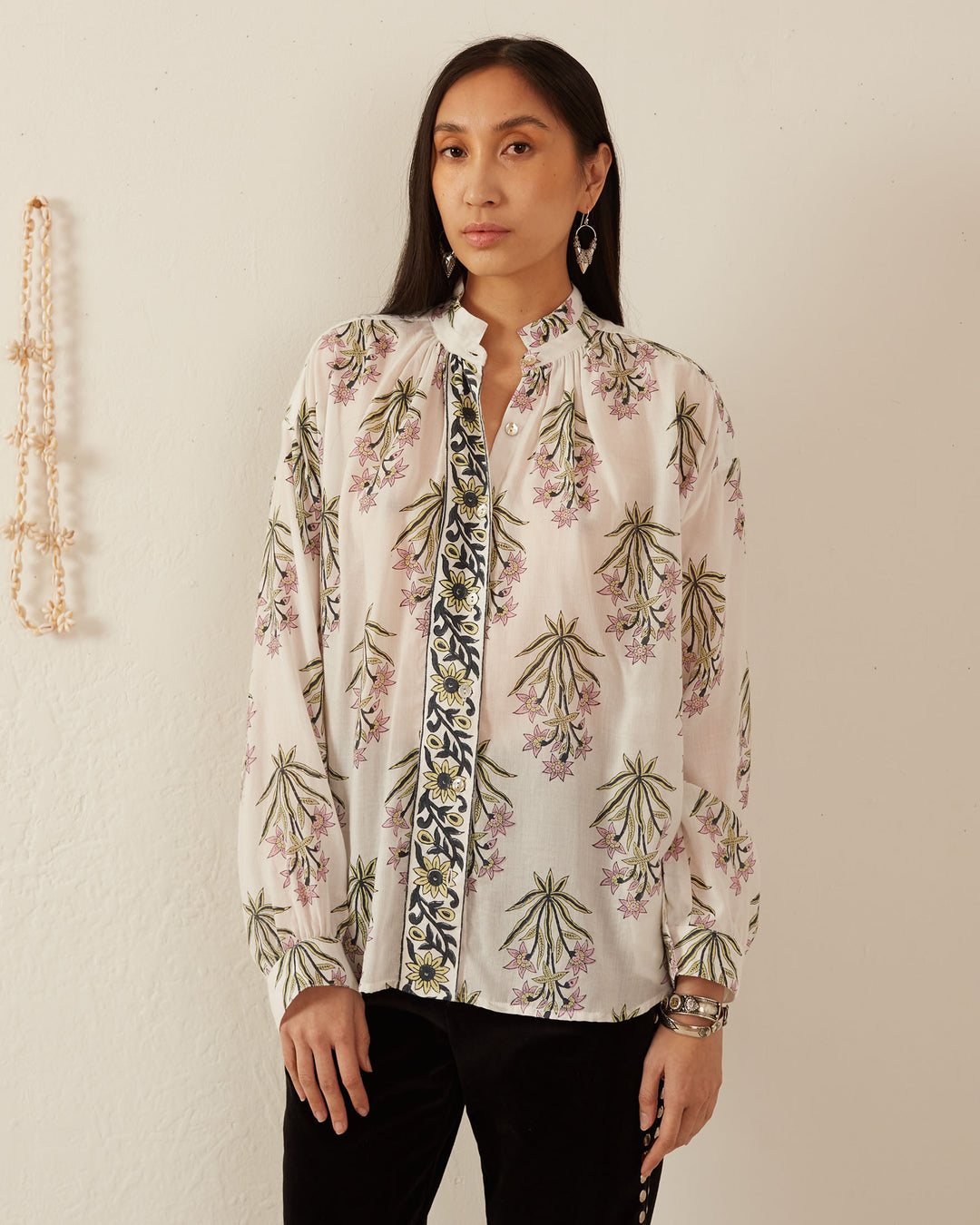 A person stands against a white wall wearing the Kiki Winter Lily Shirt, paired with black pants. The blouse features a floral pattern and is adorned with Mother of Pearl buttons. They have straight, long hair and are accessorized with earrings and bracelets.