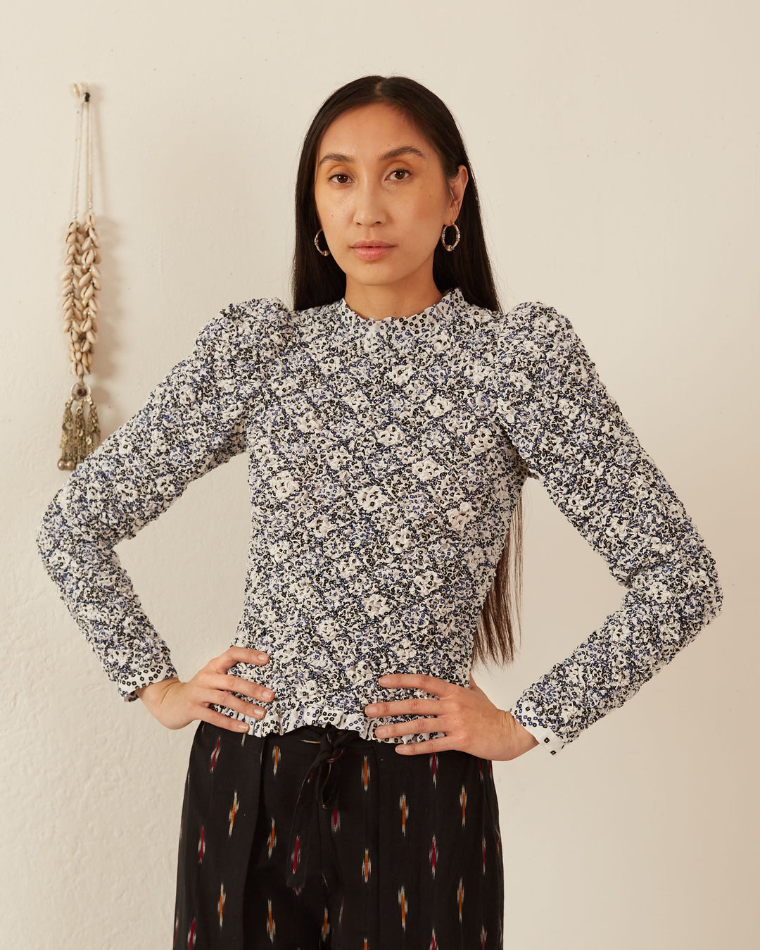 A person with long dark hair stands confidently with hands on hips, wearing the Maisie Domino Smocked Top, a floral-patterned blouse featuring a smocked bodice and ruffled armholes, paired with dark pants. The background features a light wall with a decorative hanging.