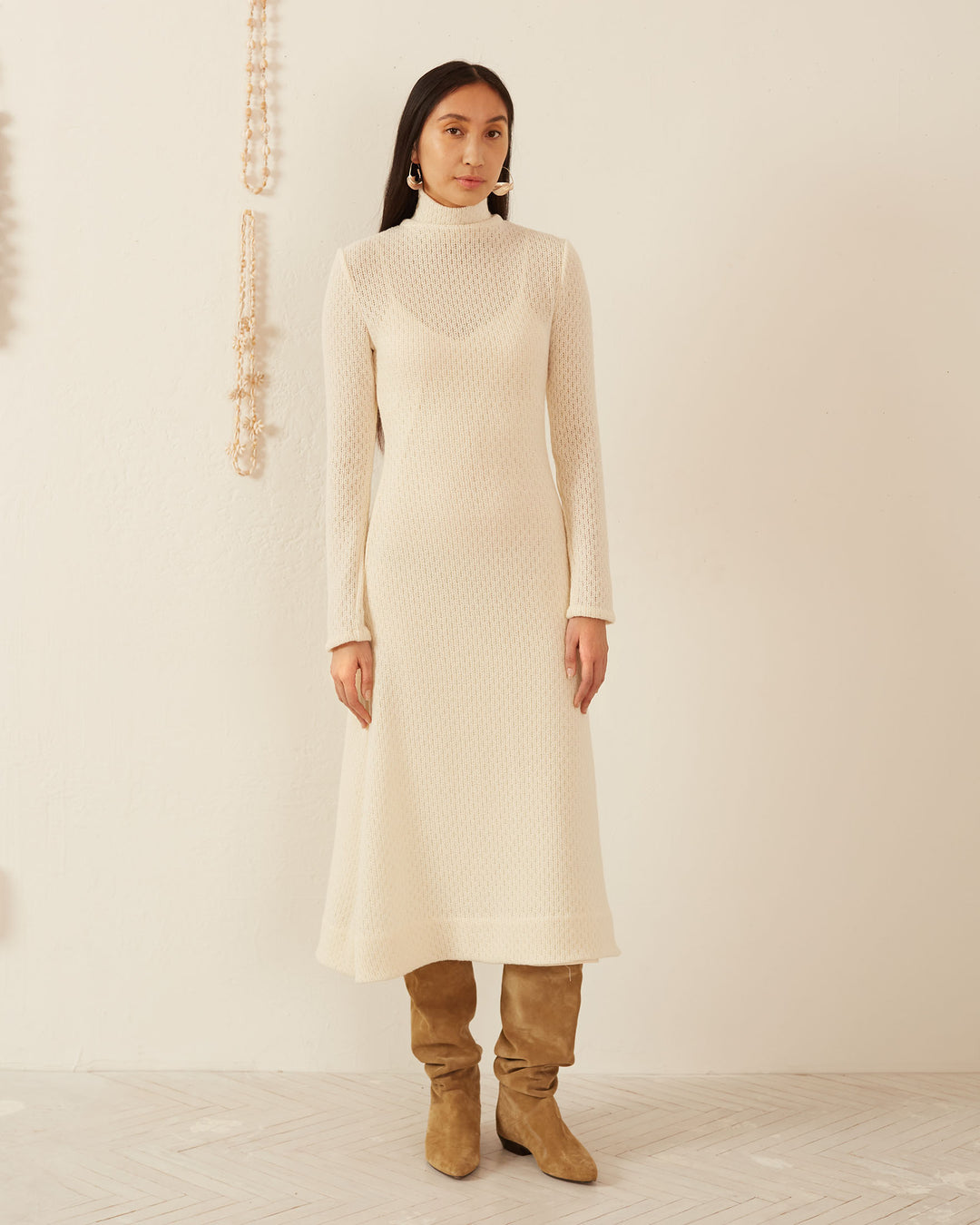 A person stands against a neutral background, elegantly dressed in the Nona Marble Knit Dress made from Italian wool, featuring a turtleneck. Complemented by tan suede boots, their ensemble includes long, dark hair and dangling earrings, with a decorative chain artistically draping on the wall behind them.