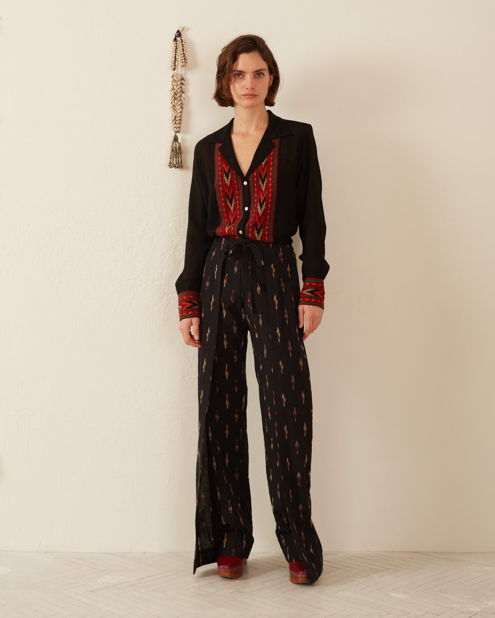 A person stands against a white wall wearing the Bonnie Etoile Wrap Pant, featuring red and white geometric patterns reminiscent of Indian ikat and an elasticated waistband. Red shoes complete the look. To the left, a decorative tassel hangs on the wall, while the floor is light-colored wood.