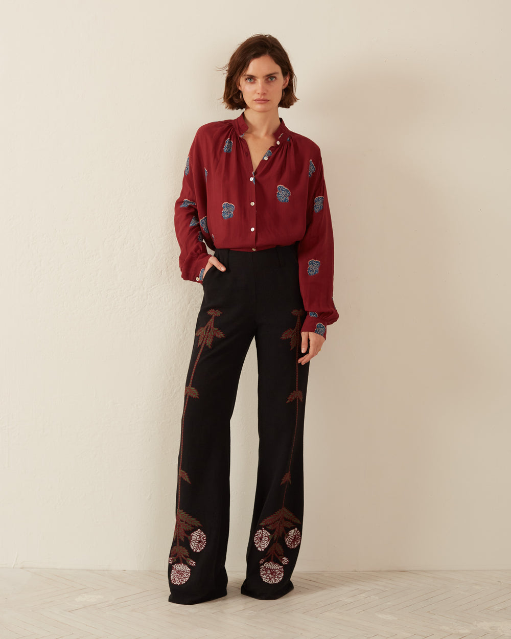 A person stands against a plain wall wearing a maroon blouse with blue and pink patterns, paired with the Charlie Black Bloom Embroidered Pant featuring hand-embroidered floral motifs. The Indian silk slub fabric adds a touch of elegance. They have short hair and a neutral expression, with hands in pockets.